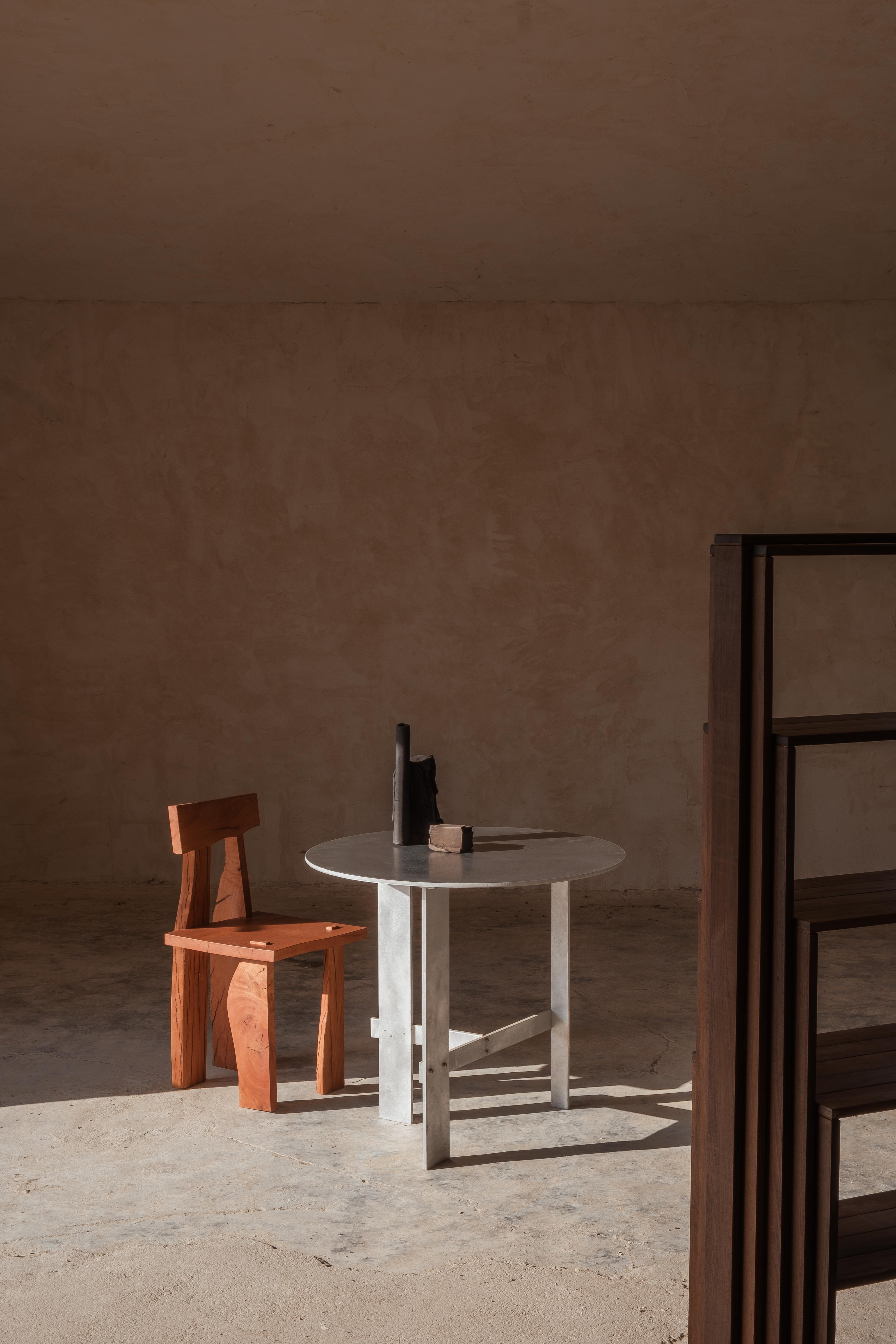 A minimalist room features a round metal table, dark geometric decor, and the sculptural burnt-orange Askew Chair by Ther. Sunlight casts bold shadows across the concrete floor and textured beige walls.