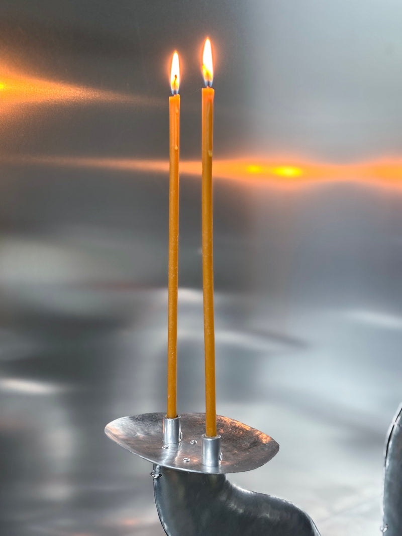 Two thin, yellow beeswax candles from the Bregje Sliepenbeek Beeswax Candles Pack of 20 burn on a modern metallic holder, their warm light contrasting beautifully with the cool, reflective silver background.