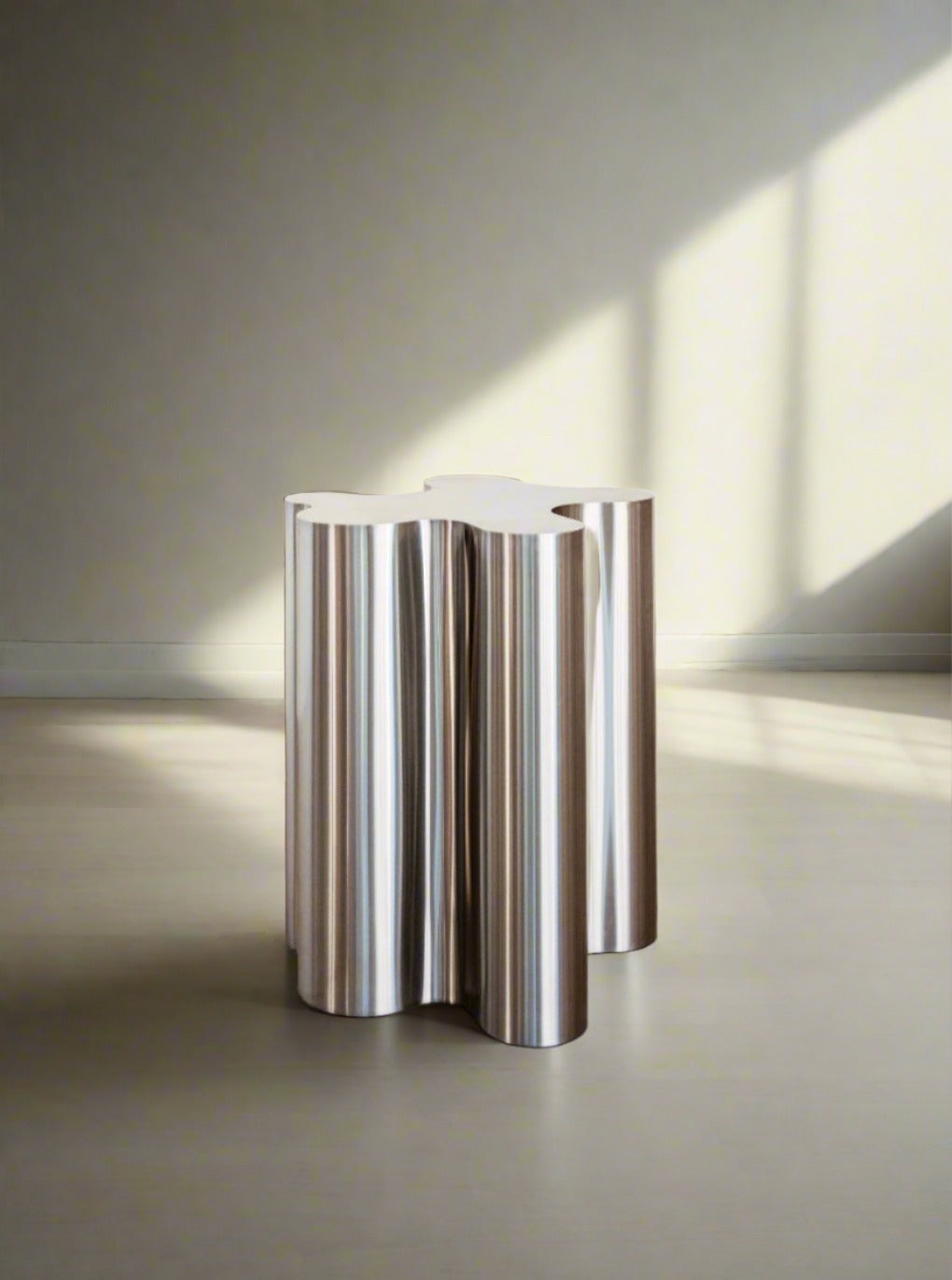 The Silver Root - Tall side table by Caia Leifsdotter, with its sculptural steel and wavy four-lobed design, stands in a minimal room as soft light streams through a window, casting shadows on the floor and wall.