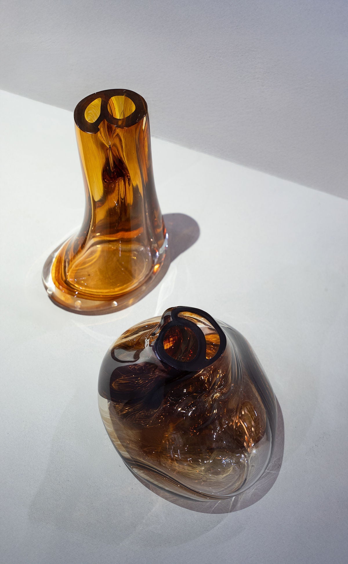 Two abstract, amber-colored HOMA Elara Vase IV pieces with organic shapes rest on a light surface, casting soft shadows. Each mouth-blown vase has two round openings at the top and a glossy, reflective finish.