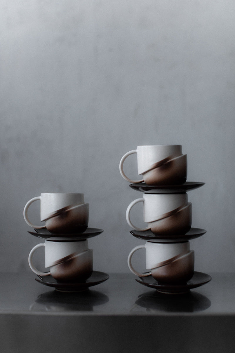 Six Art Deco Ceramic cups with saucers by Out For Lunch are stacked in two columns on a gray surface. Featuring a modern brown-and-white gradient, this set captures the vintage charm of classic serveware.