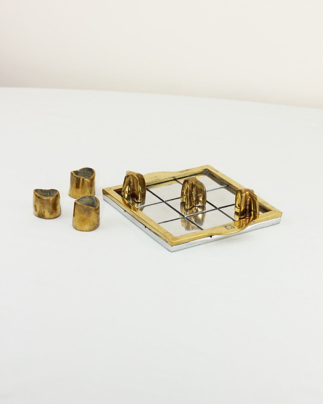 TRES EN RAYA BRUTALISTA — angled view of brass game pieces on white surface