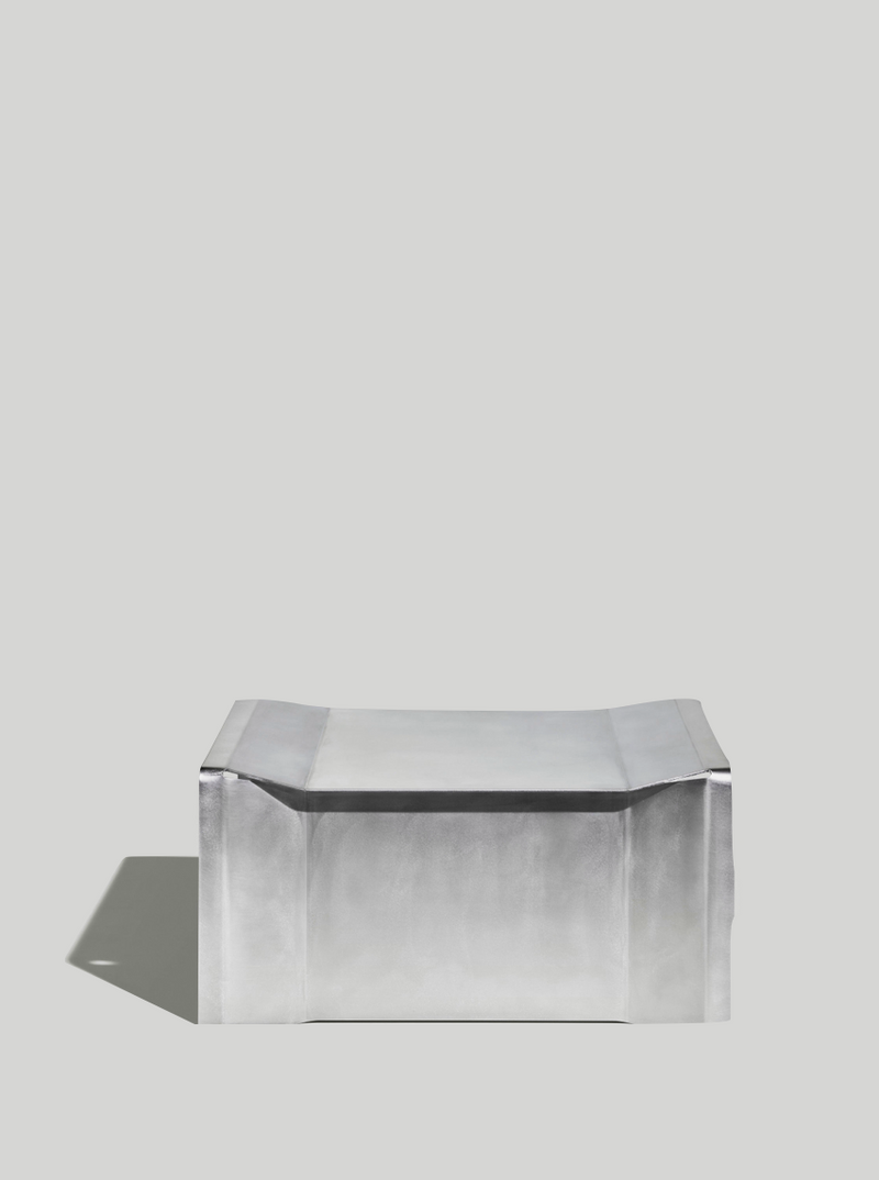 The Camber Coffee Table by Paul Coenen is a sleek, modern rectangular metallic table with slightly flared top edges and an angular design, casting a soft shadow on a plain light gray background.
