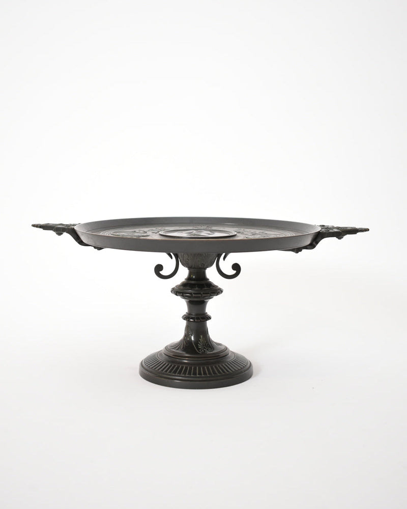 The Barbedienne Style Pedestal Cup, XIXth c. by Barracuda Interiors is a decorative black metal tray with ornate handles and an engraved round surface, styled with an antique patina and displayed against a plain white background.