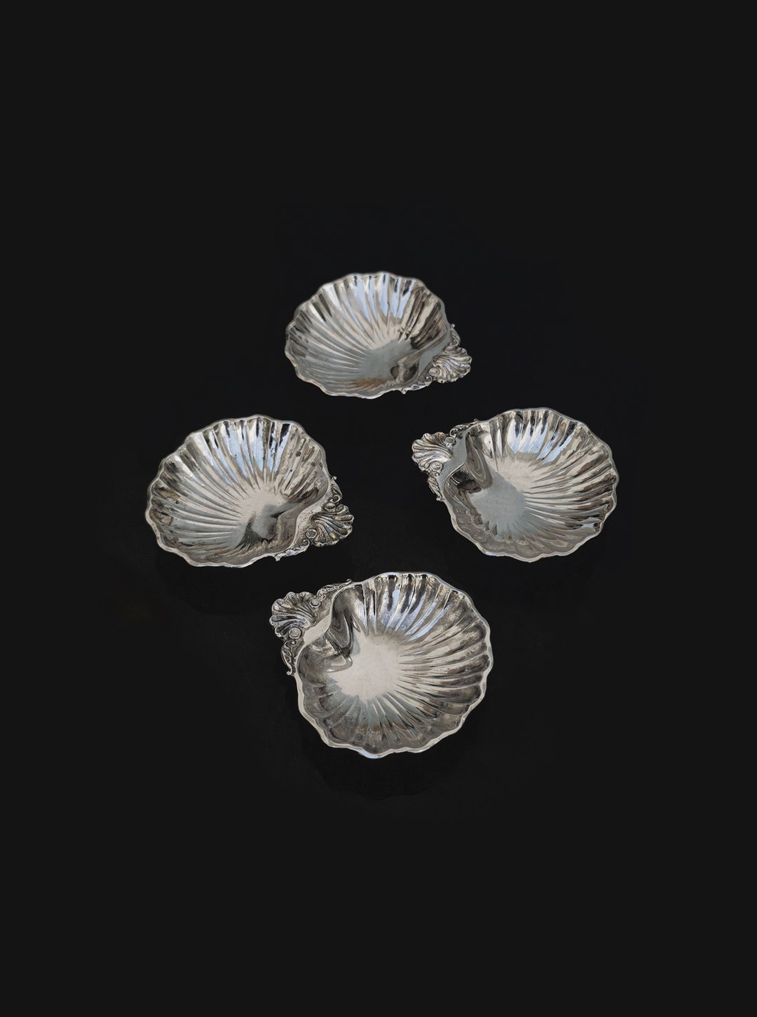 The Les Objoies Set of 4 small shell vide poches features ornate silver-plated dishes with decorative handles, elegantly arranged in a diamond pattern on black for a striking decorative display.