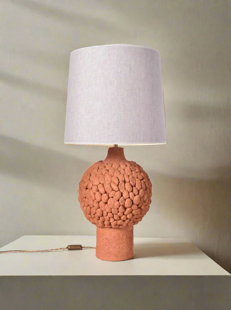 The "Melides" Brown Glazed Terracota Lamp by Barracuda Interiors features a textured round base and a white fabric shade, placed on a light surface against a neutral wall, with the cord and switch visible.