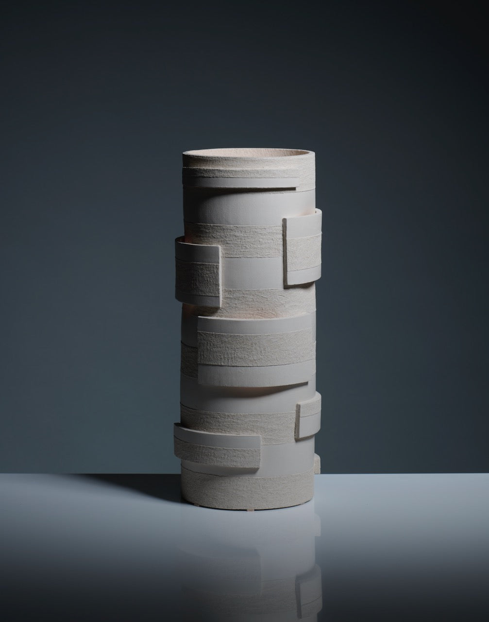 The "Bricks" Table Lamp in ceramic by Katia Langeard features a tall, cylindrical shape with matte off-white finish and layered rectangular forms, shown on a glossy surface against a dark gradient—ideal for hallway accent lighting.