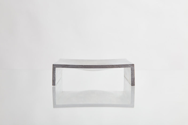 A minimalist, dark rectangular bench or table with a curved seat features the LESORR Square Aluminum Vide-Poche as a small object holder. It sits on a reflective white surface against a plain white background.