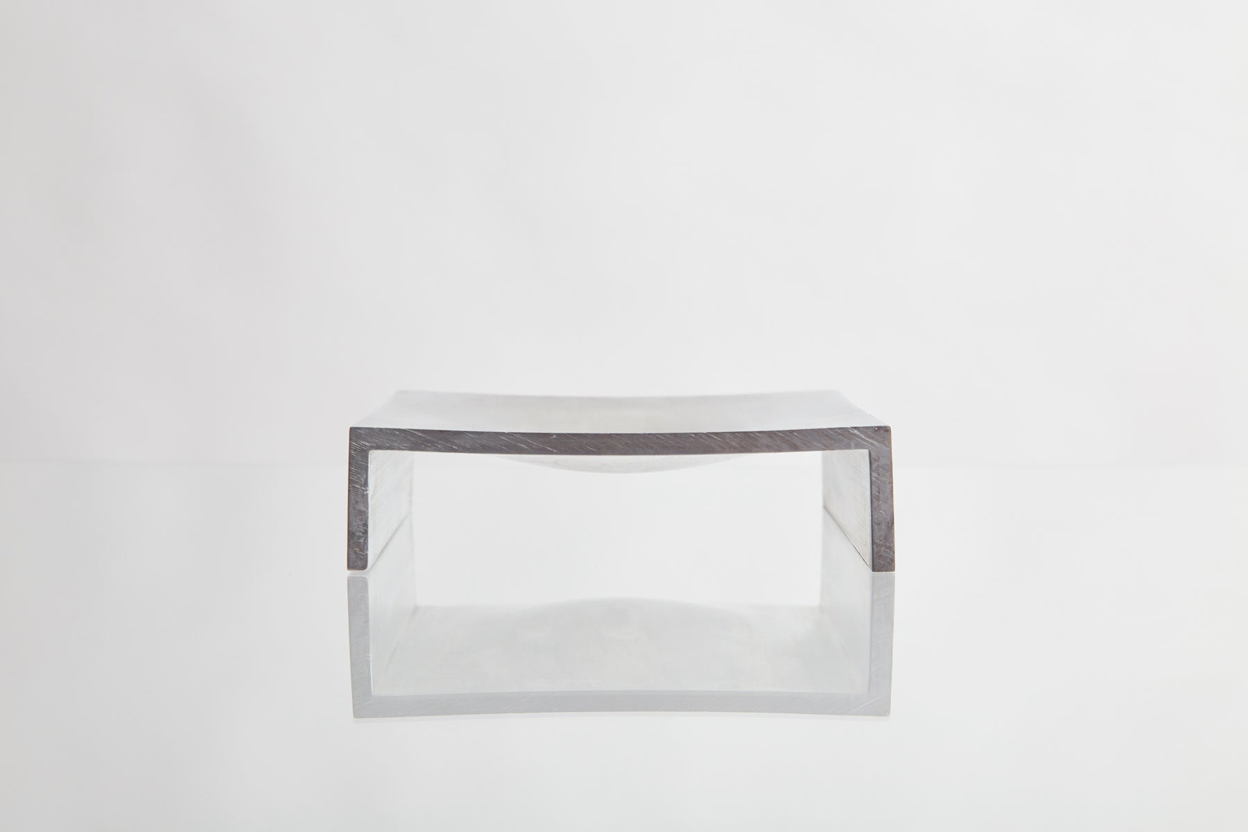 A minimalist, dark rectangular bench or table with a curved seat features the LESORR Square Aluminum Vide-Poche as a small object holder. It sits on a reflective white surface against a plain white background.