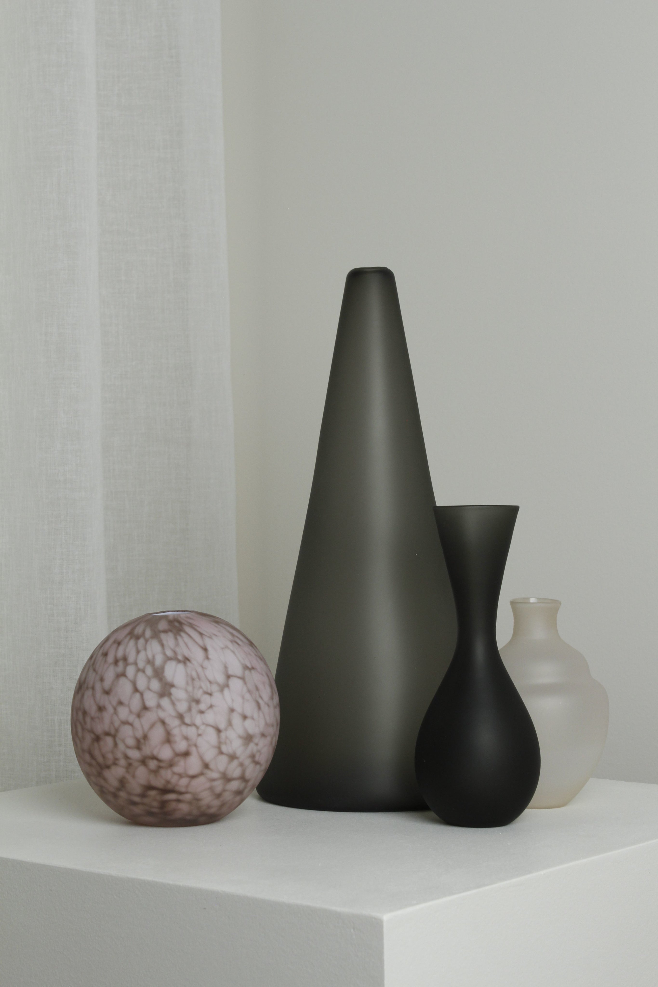 The Medium Vase in Smoke by Verre d'Onge, with its sleek silhouette, sits stylishly on a white pedestal against a neutral backdrop.