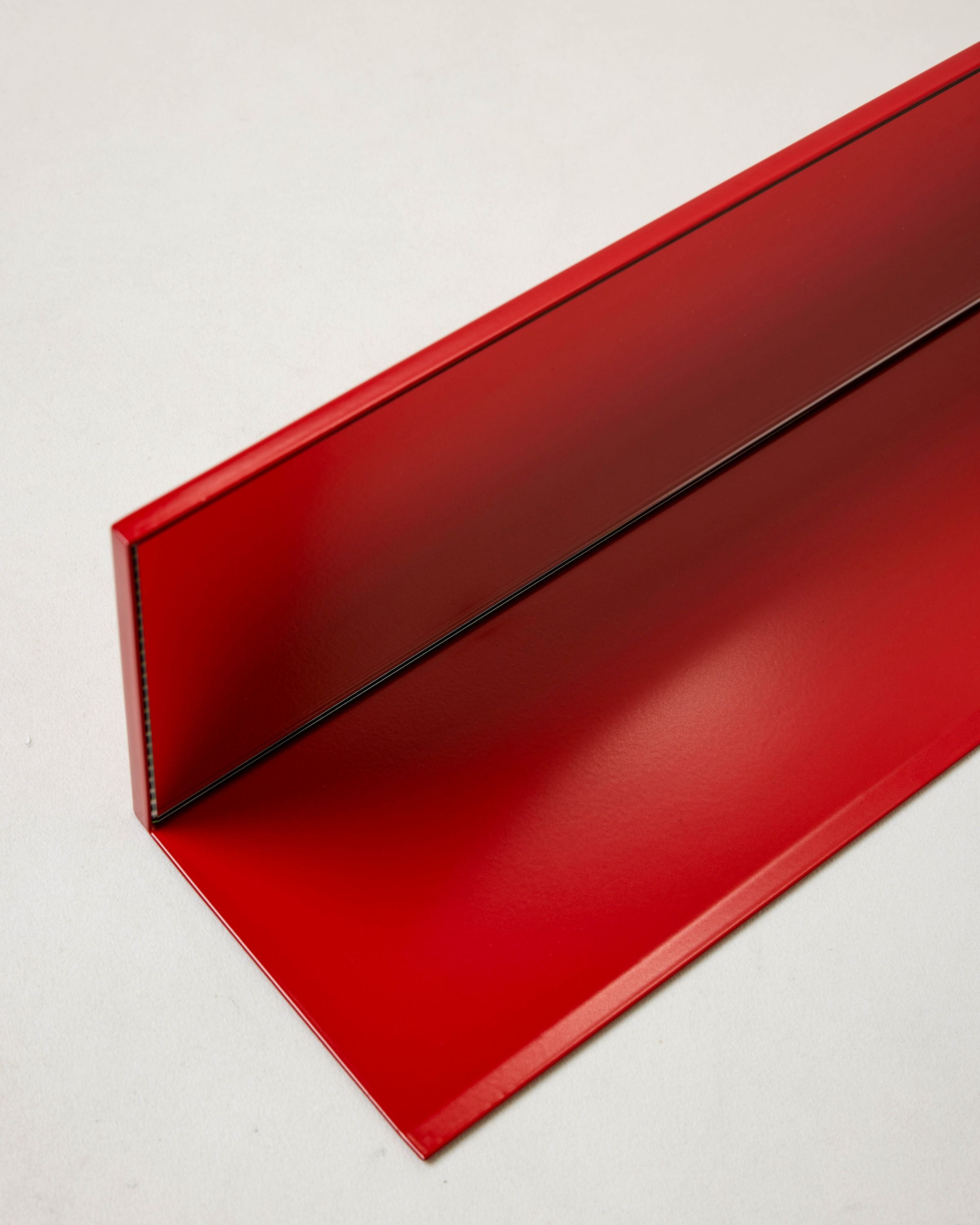 The Dot Objects Mirror Steel Shelf, an L-shaped red metal bracket ideal for customizable shelving, is shown resting on a light-colored surface in an angled top-down shot.