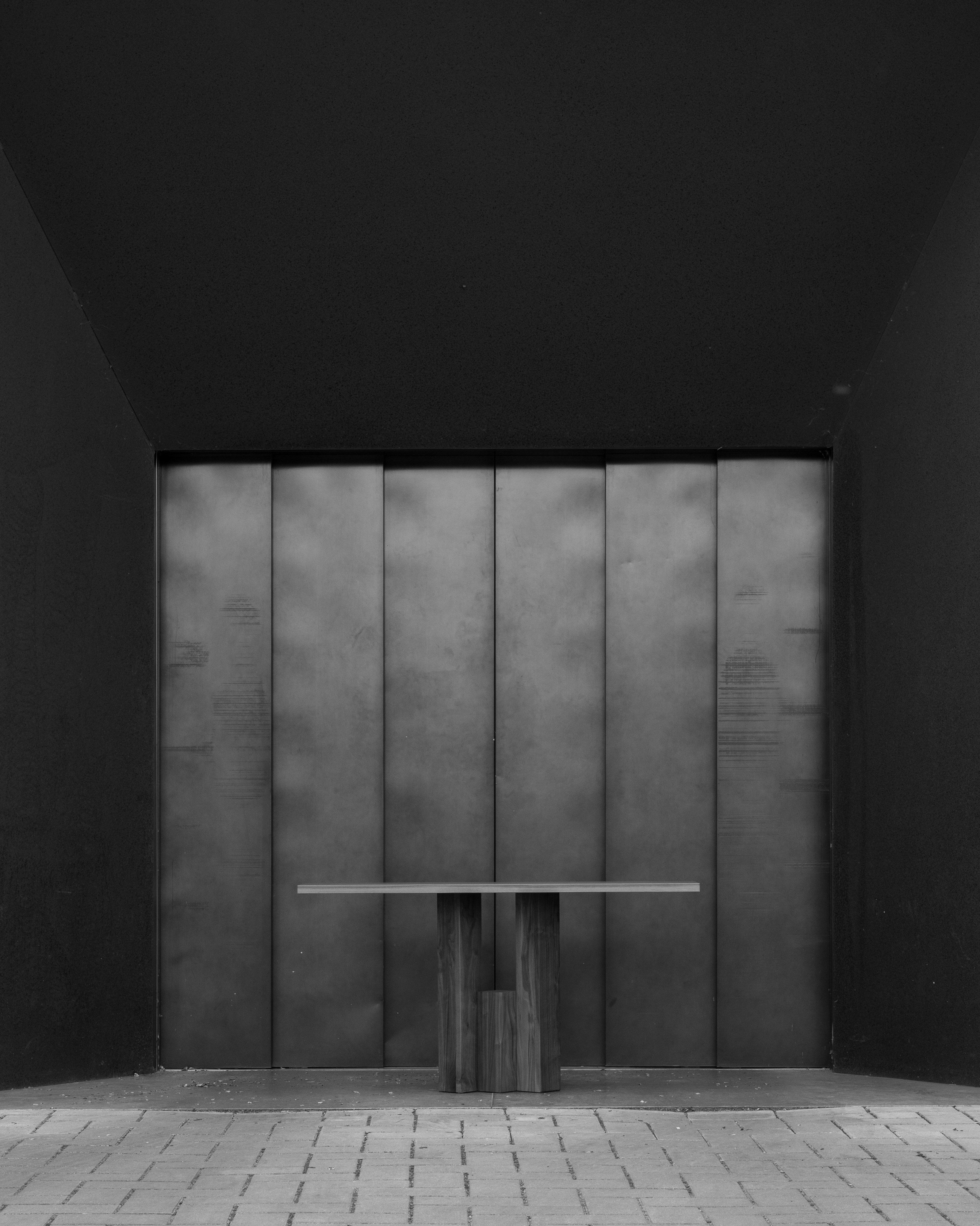 A minimalist black and white photo features the Mokko Fenestra Console, a customizable bench with a rectangular seat and wooden supports, placed in front of a flat metallic door styled with Brick Expressionism, all on tiled flooring framed by dark walls.