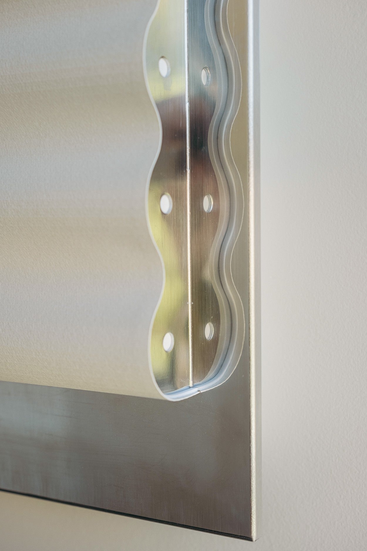 Close-up of the "Frame" Mirroir M by Violaine d'Harcourt, showing its metal corner bracket with a wavy edge and round holes attached to a flat surface. The softly blurred background highlights its shiny, minimalist design.