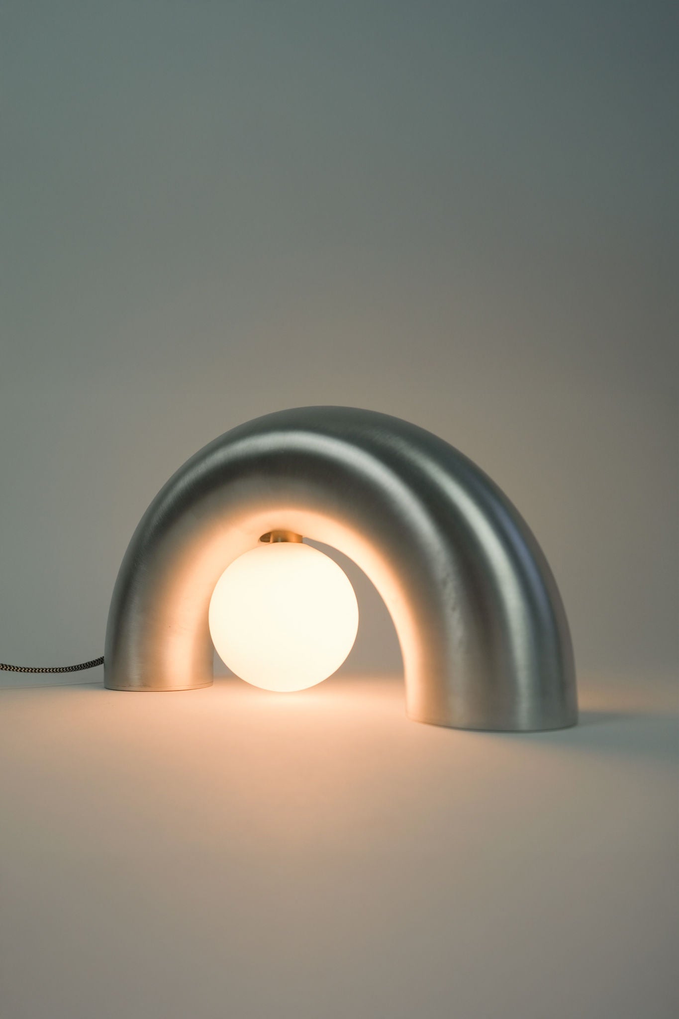 The Arcora Underglow - Aluminum Arc Lamp by Heako Studio features a brushed metallic arc with a glowing white bulb beneath, adding a calming, modern touch to any neutral space.