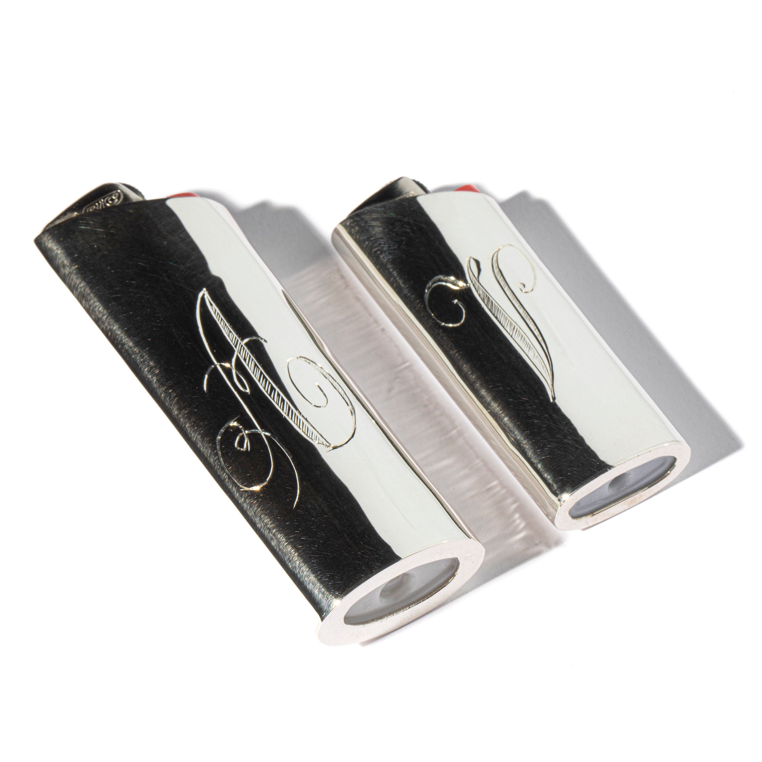 Two Pedro Ventura Monogram Lighter Case Maxi Size pieces, crafted in sterling silver with engraved swirling designs and holding standard red-top lighters, are displayed on a white background with shadows to the right.