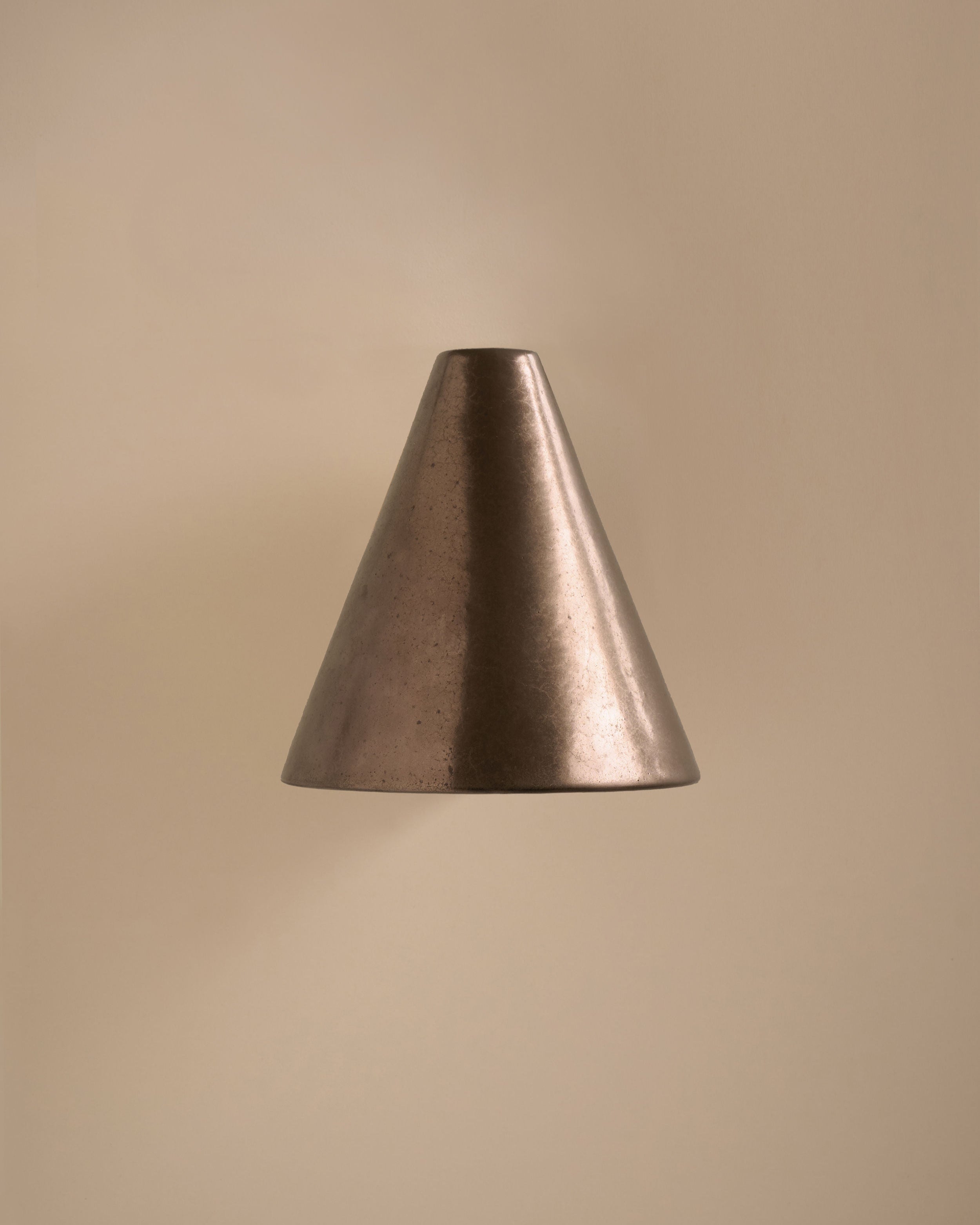 The 'Esoteric' Wall Light by Mariza Galani features a handcrafted ceramic, metallic cone design with a smooth finish, mounted on a beige wall. It casts soft light below, creating a warm and minimalistic ambiance.