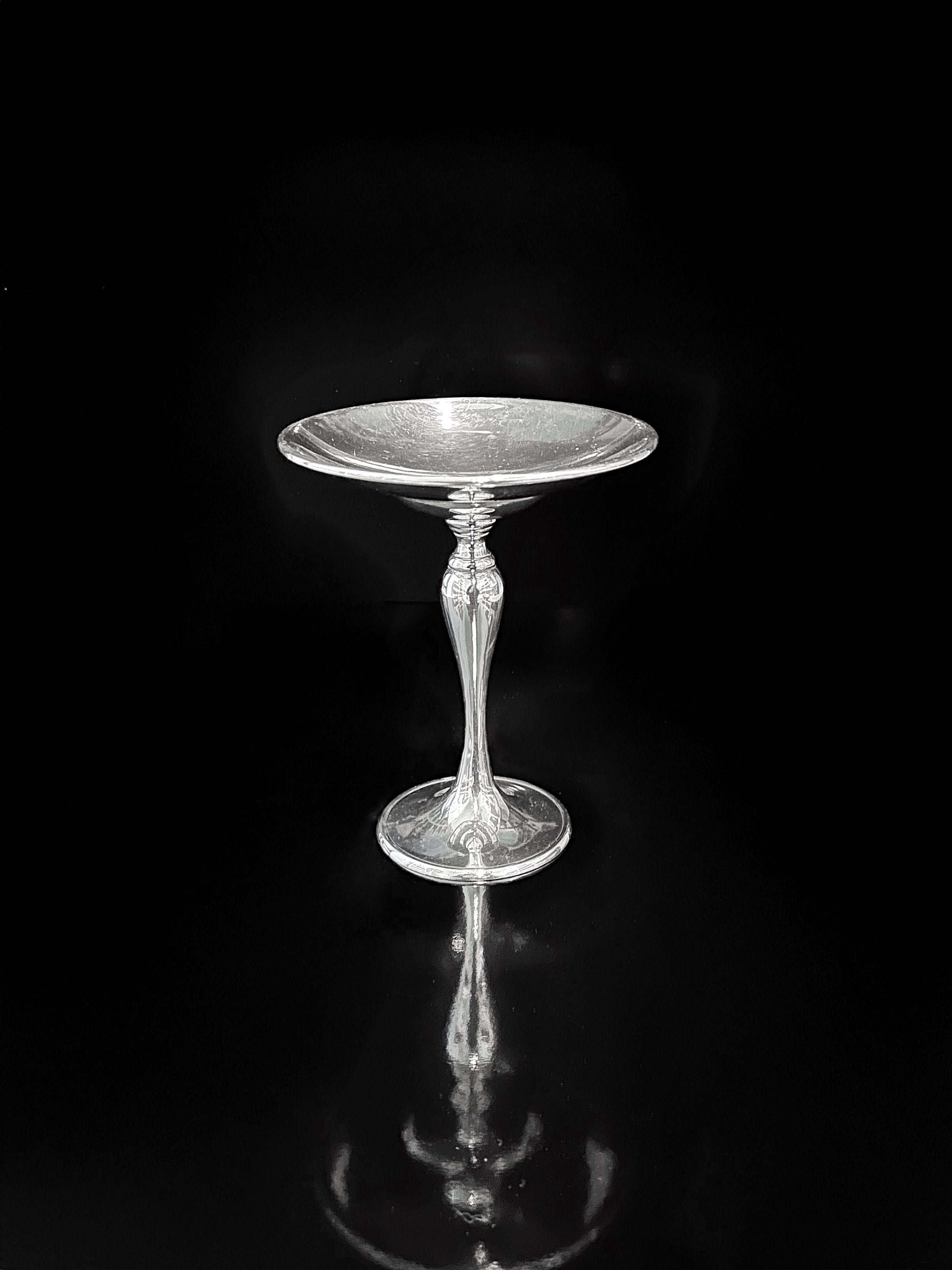 A Dodo Vintage Heavy Silver-Plated Foot Bowl stands centered on a glossy black surface, its vintage silver finish and elegant stem casting a subtle reflection.