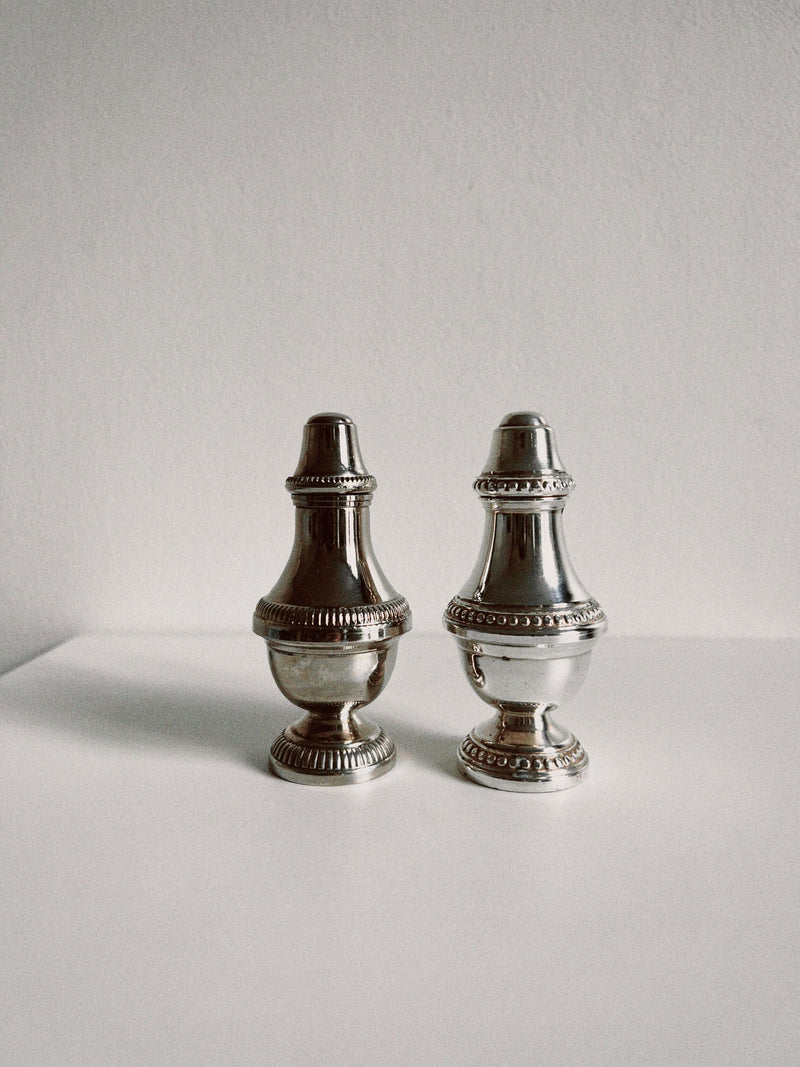 The Manushi Silver Salt and Pepper Shakers, with ornate detailing, are displayed side by side on a white surface against a light gray background, highlighting their classic elegance.