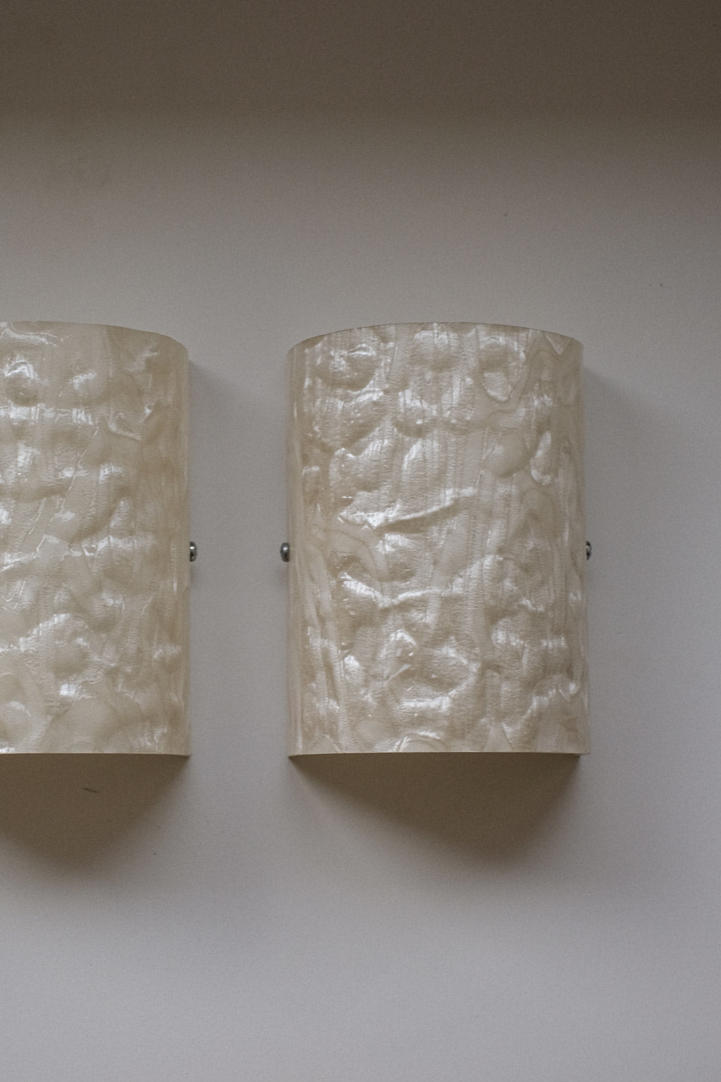 The Set of 2 Mother-of-pearl Wall Lamps by septembre studios features textured, cylindrical shades with a glossy, wavy pattern, mounted on a light wall and evoking the vintage charm of 1980s lighting design.