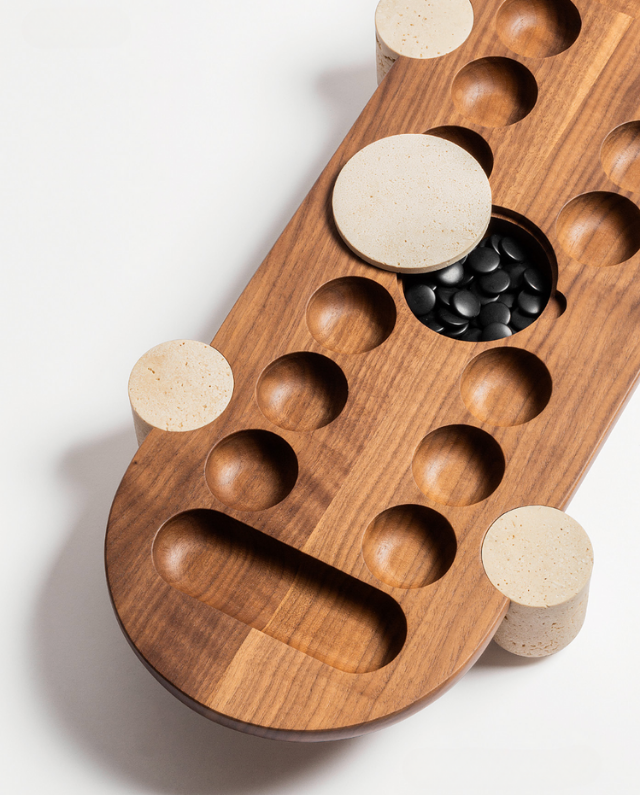 The GIOBAGNARA MOCAMBO Wari Game Set features a solid walnut board with carved pits, round stone cups on each corner, and black playing stones, all elegantly displayed on a white background.