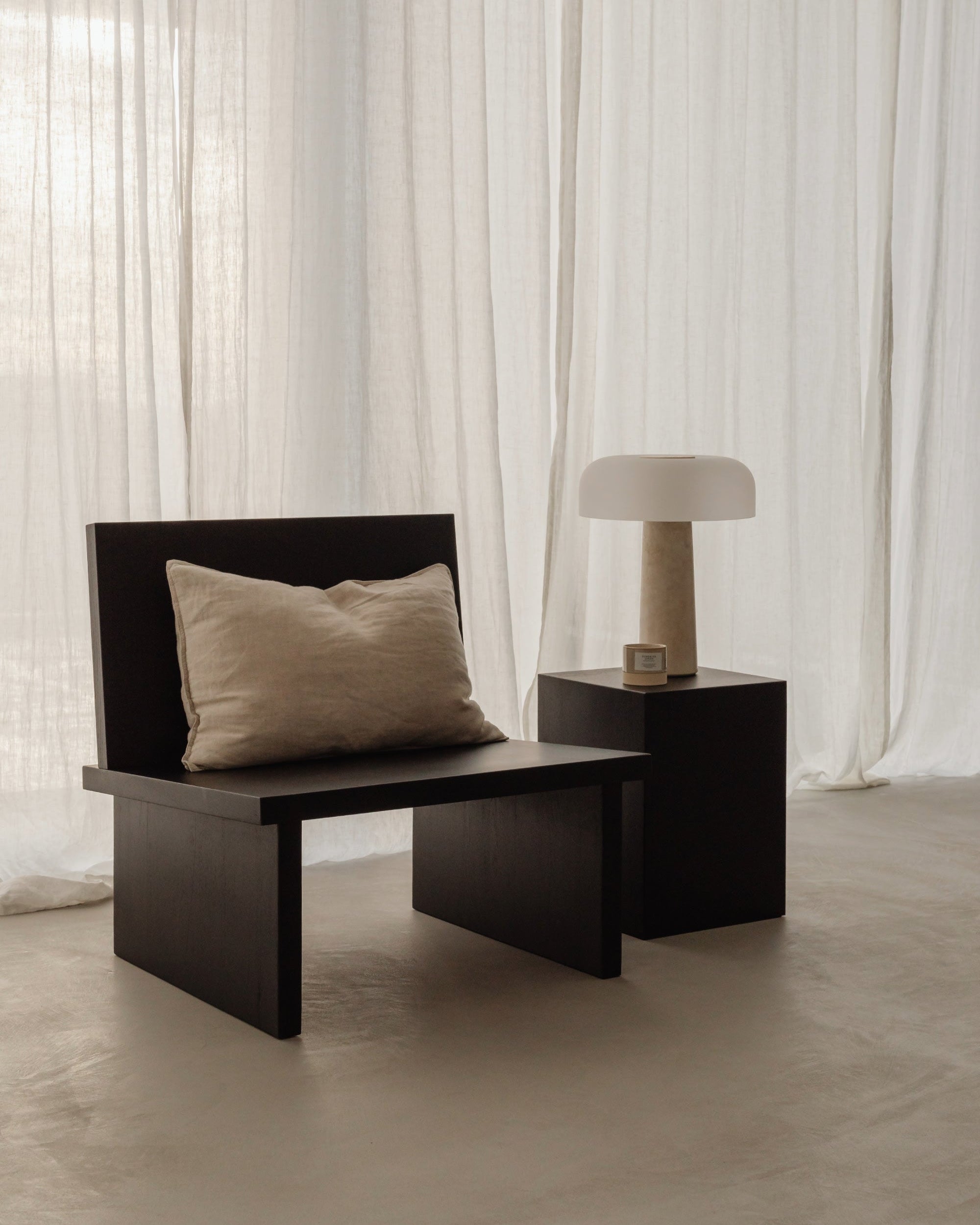 A minimalist scene featuring the ZEBU Chair by un'common, with black wood and a beige cushion, placed next to a square black side table holding a modern lamp, set against sheer white curtains and a light-colored floor.