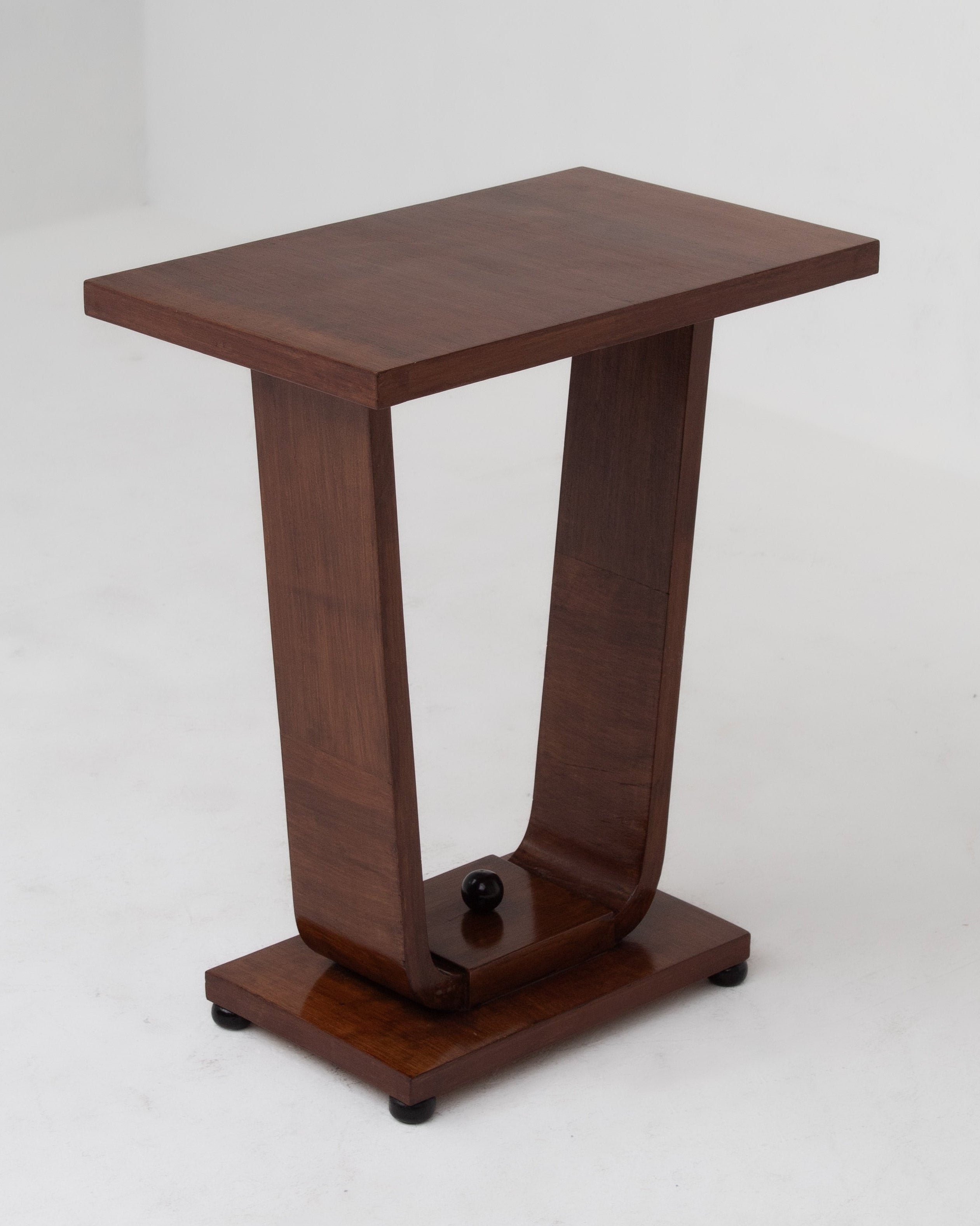 The Malata Antwerp Low Art Deco Side Table features a sleek, modern wooden design with a narrow rectangular top, central curved support, rectangular base, and a small black decorative sphere. Shown against a plain white background.