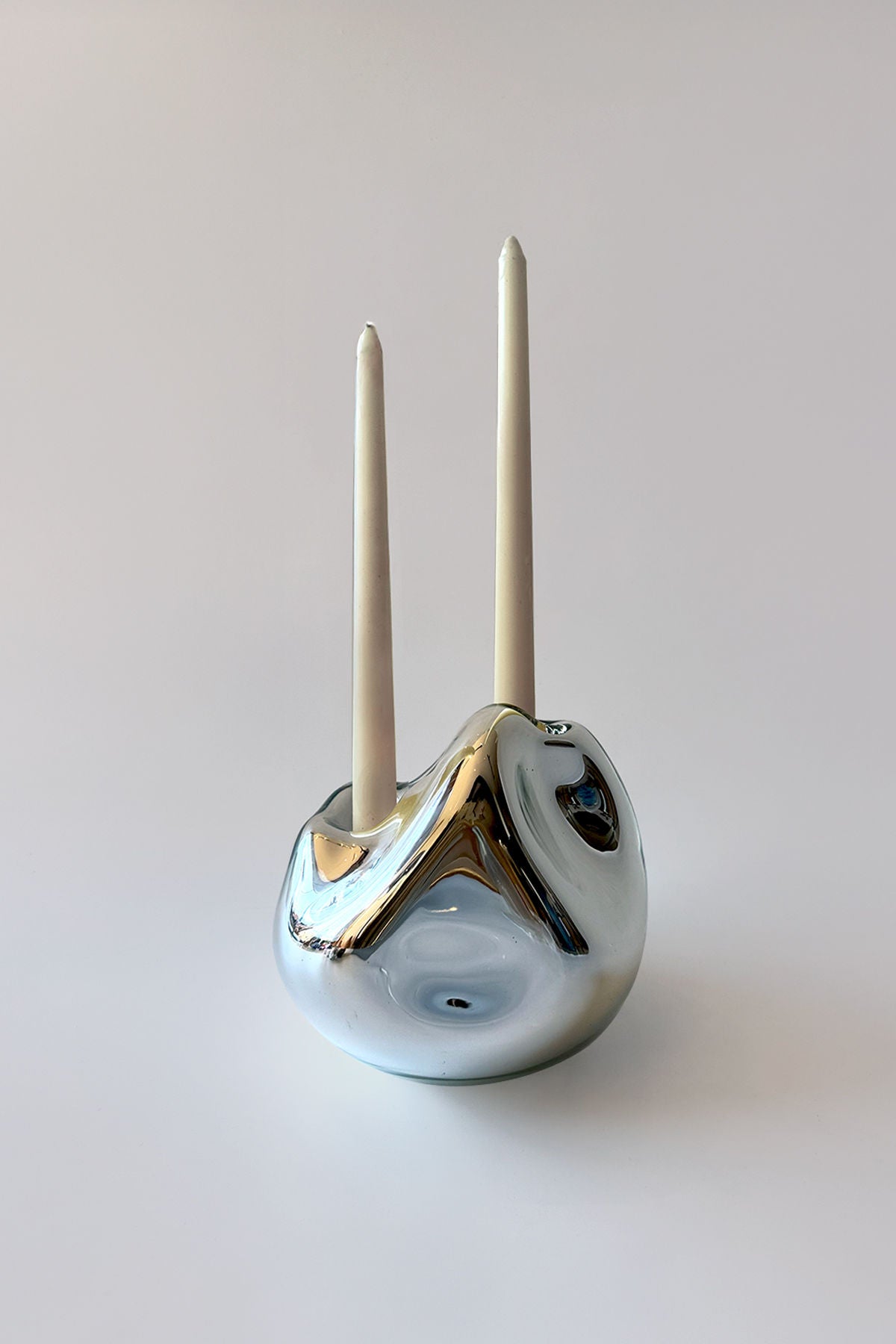 The HOMA Lumi Candle Holder V is a shiny, abstract silver piece with a mirroring finish and organic, rounded form, displaying two tall white taper candles against a plain light background.