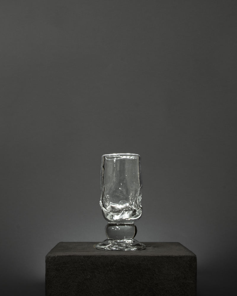An empty Organic Footed Table Glass by Szkło Studio, hand-blown with a textured surface, stands on a dark stone pedestal against a plain, dark gray background.