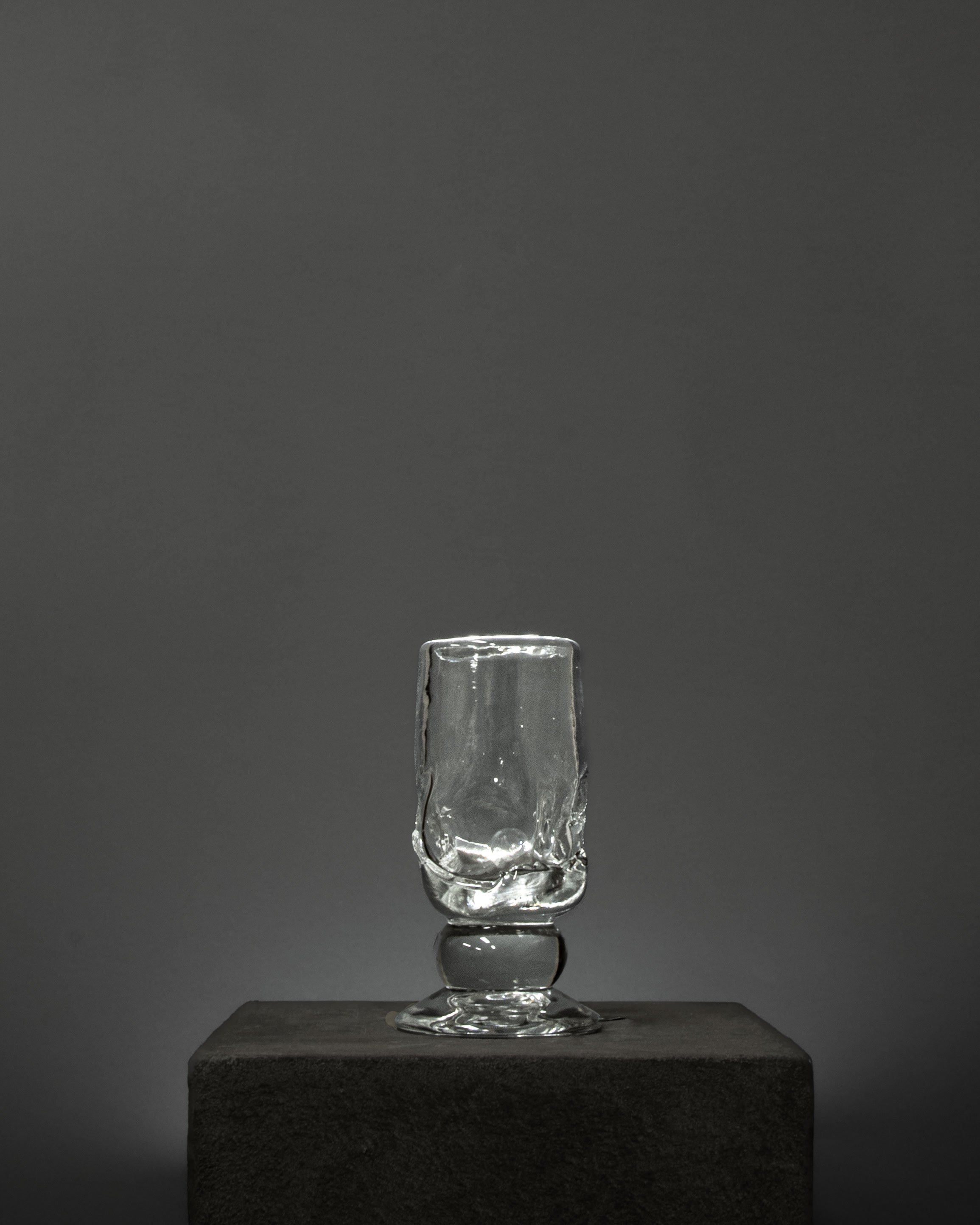 An empty Organic Footed Table Glass by Szkło Studio, hand-blown with a textured surface, stands on a dark stone pedestal against a plain, dark gray background.