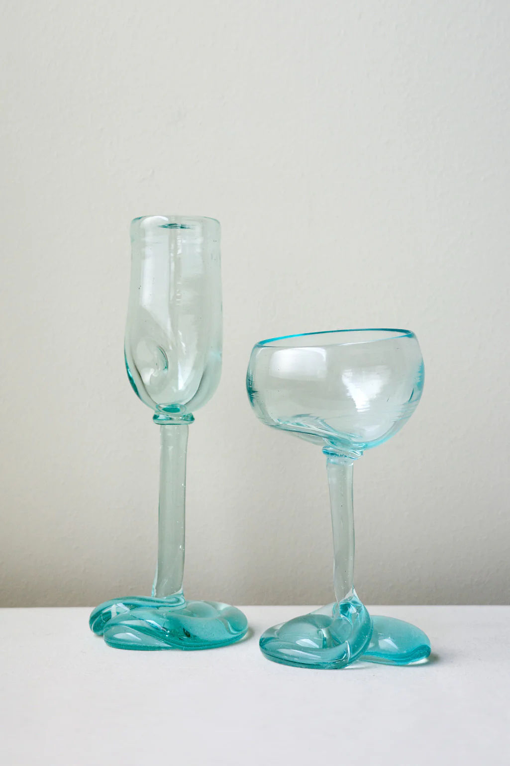 Two clear, hand-blown Recycled Flute Glasses by Completedworks feature wavy stems and sculptural melted designs, with one tall flute-style glass and one shorter bowl, embodying artistic recycled glass craftsmanship.