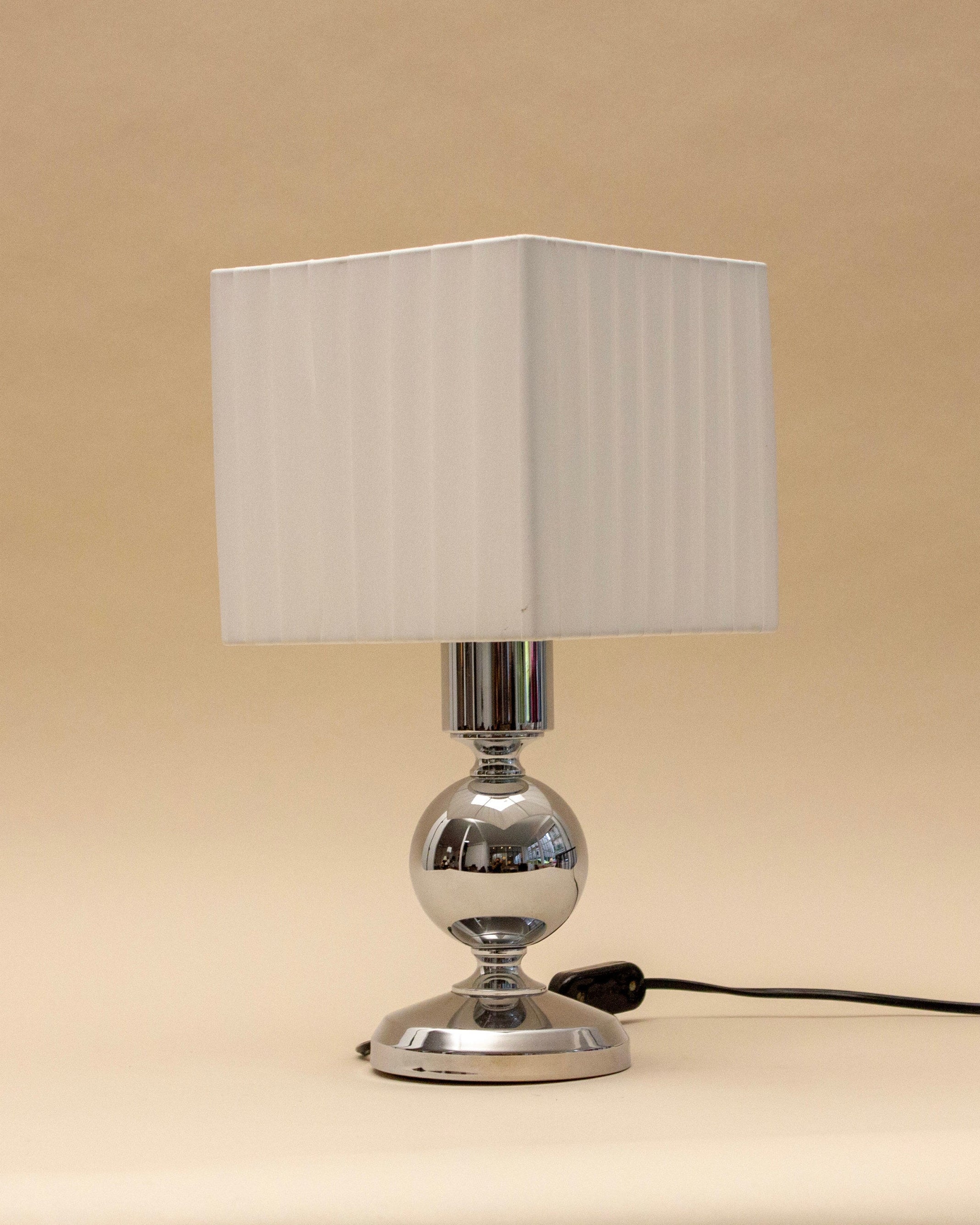 The SÒLER ALMADA WHITE LAMP features a shiny metal base and a square, white pleated shade. Plugged in against a plain beige background, this modern table lamp adds elegance to any space.