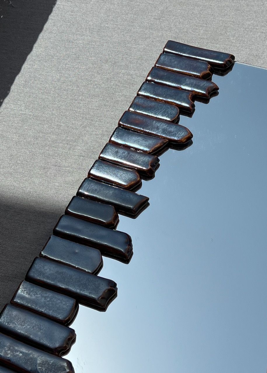 A close-up of the Cliff Collection Contemporary Mirror by Katia Langeard, featuring an irregular wavy black ceramic frame on gray fabric, reflects the sky and casts soft shadows.