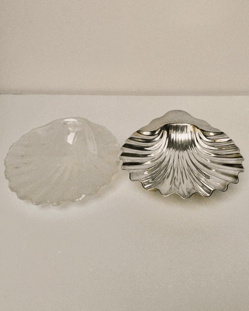 The Manushi Silver Shell Shaped Dish is displayed alongside a glass shell dish on a white surface with a plain background.