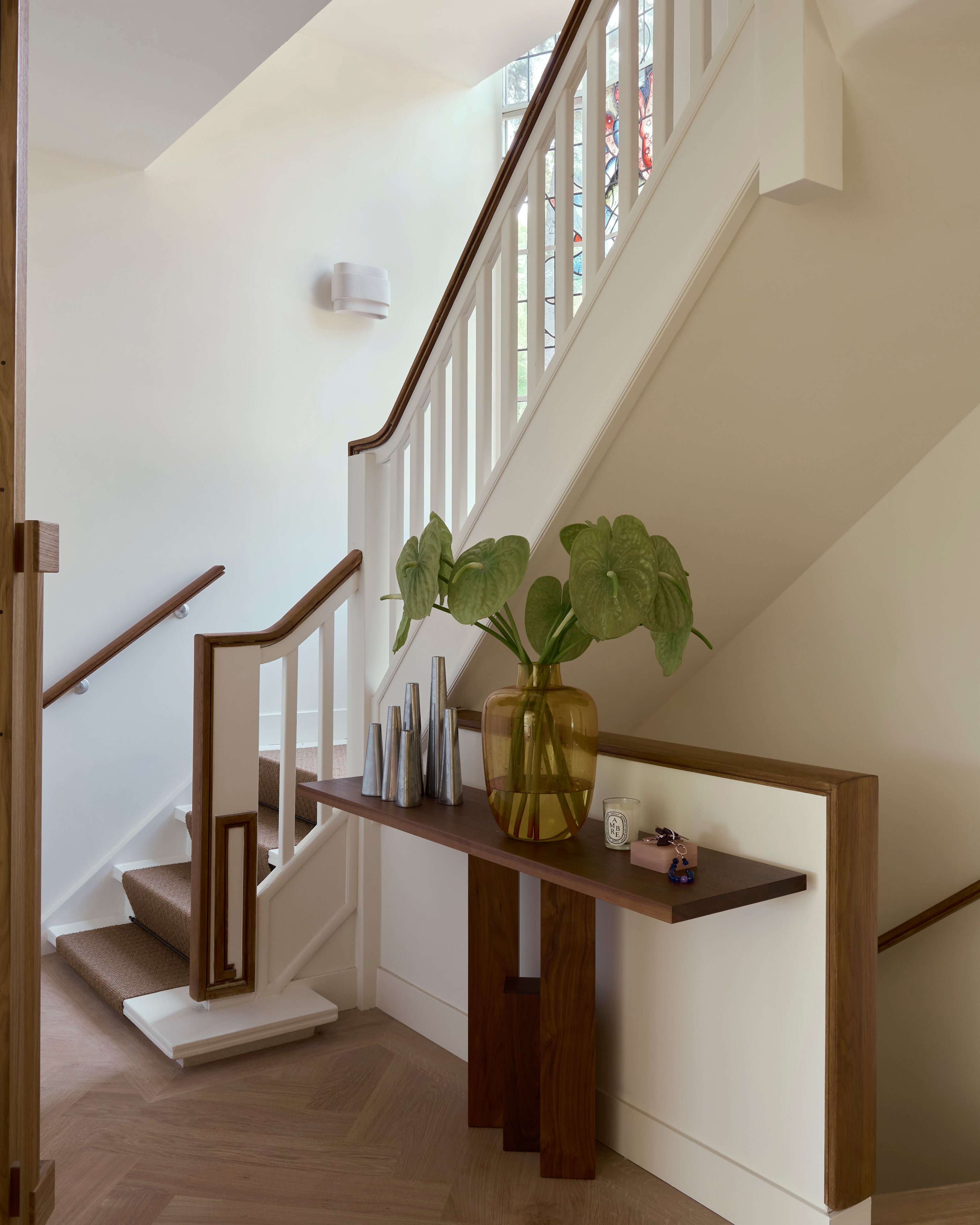 A modern hallway with a wooden staircase, white railings, and the Mokko Fenestra Console displaying a large yellow vase with green leafy plants, books, and decor pieces beneath the stairs.