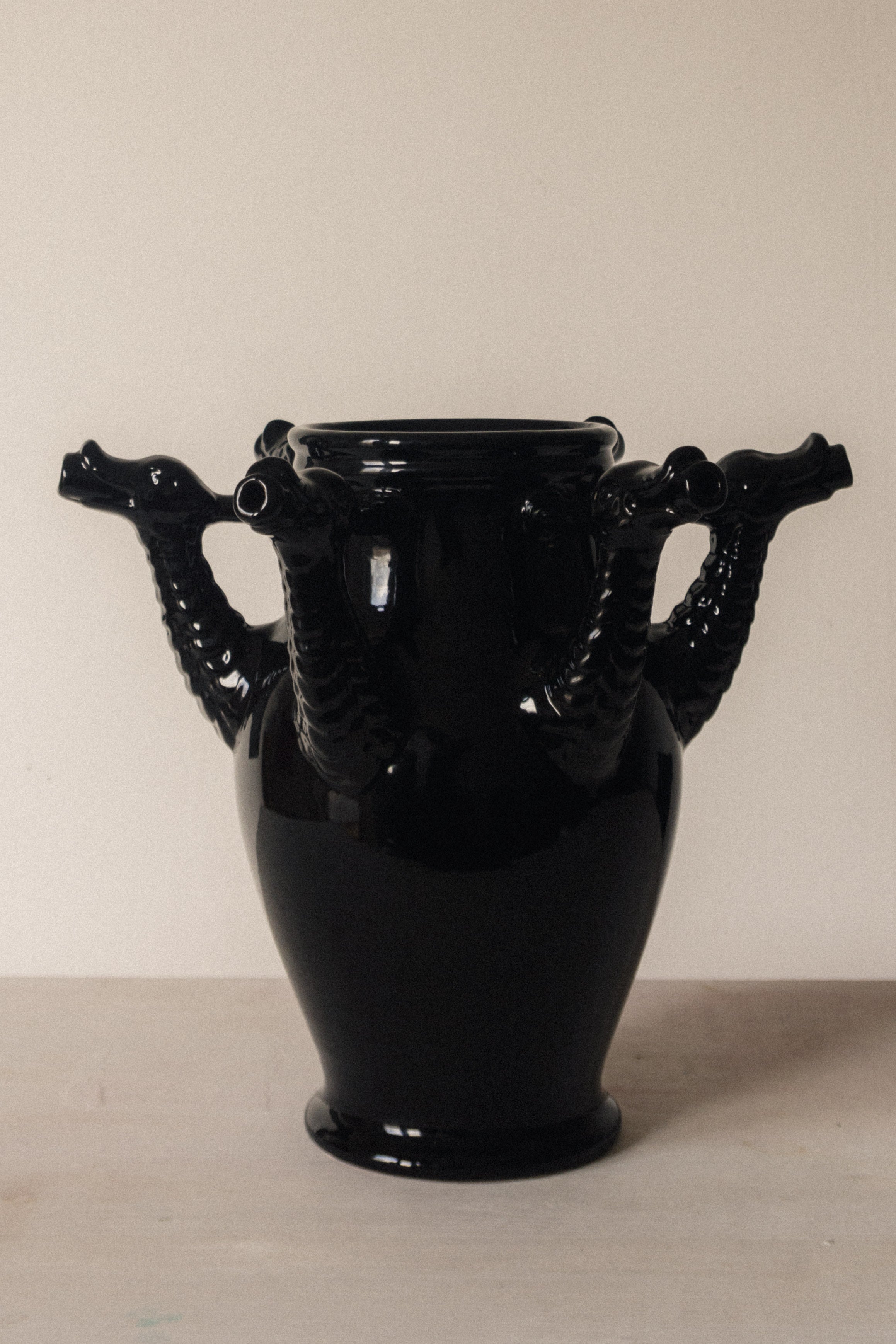 The Drago Bianco by COSEINCORSO is a glossy black minimalist ceramic vase, handmade in Italy, featuring four sculpted dragon head handles symmetrically arranged against a plain, light background.
