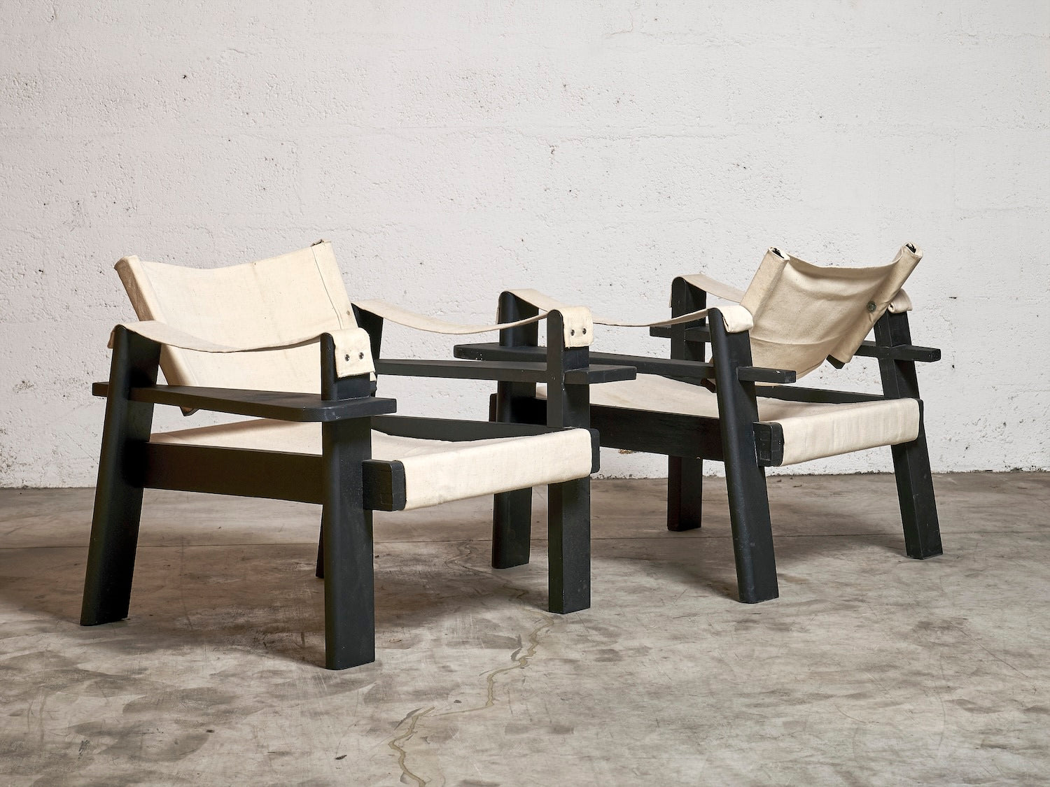 A pair of "Low Chairs in the Taste of Pierre Dariel" by Harold Mollet, featuring black painted wood frames and off-white fabric seats and backs, are positioned side by side on a concrete floor against a plain white wall.