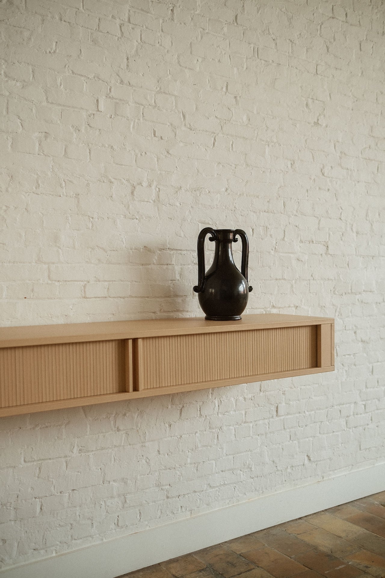 The Fiasca - Minimalist Vase in Ceramic by COSEINCORSO, a black two-handled piece, rests on a light wooden floating shelf against a white painted brick wall, with warm-toned floor tiles partially visible.