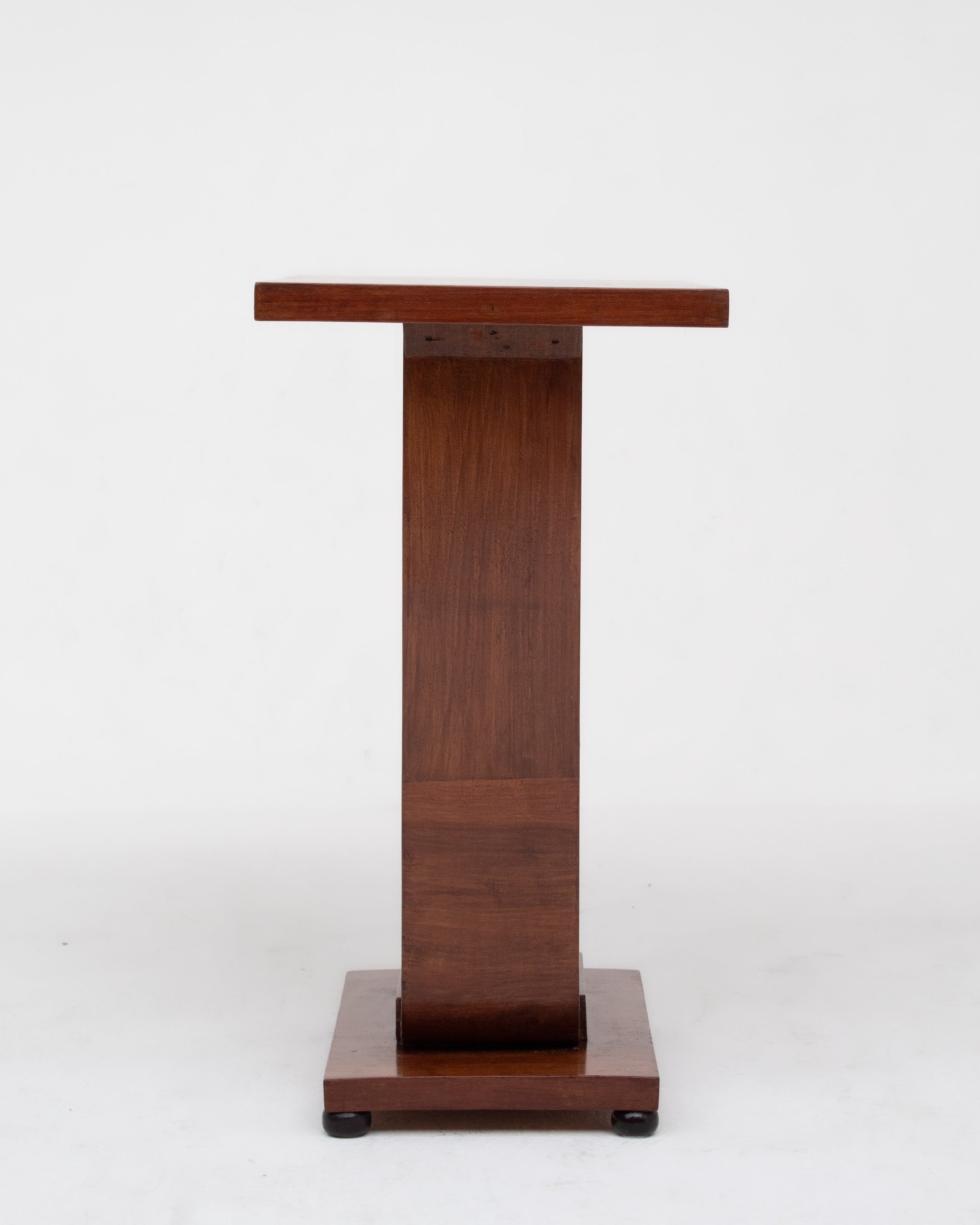 The Malata Antwerp Low Art Deco Side Table features a simple, rectangular wooden design with a narrow profile and square base with small wheels, resembling a vintage podium, shown against a plain white background.