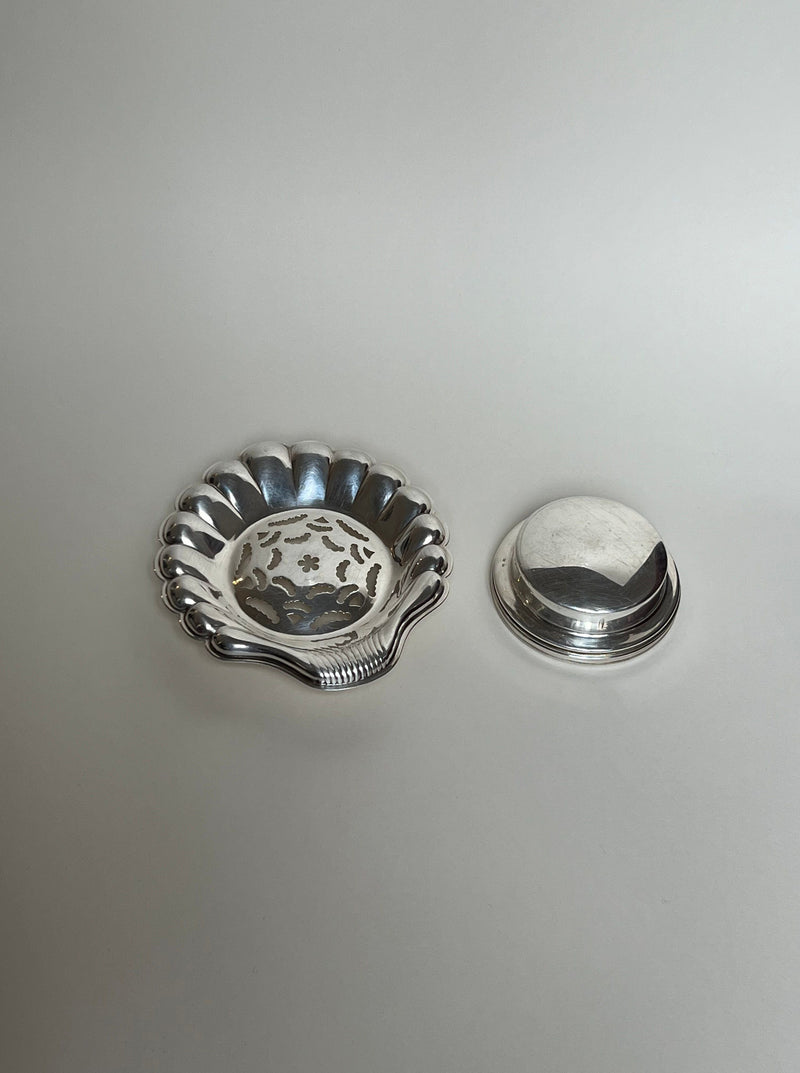 On a light background: Manushi's Vintage Shell Shaped Butter Curler with decorative cutouts on the left, and a smooth, round silver lid or vintage utensil base on the right.