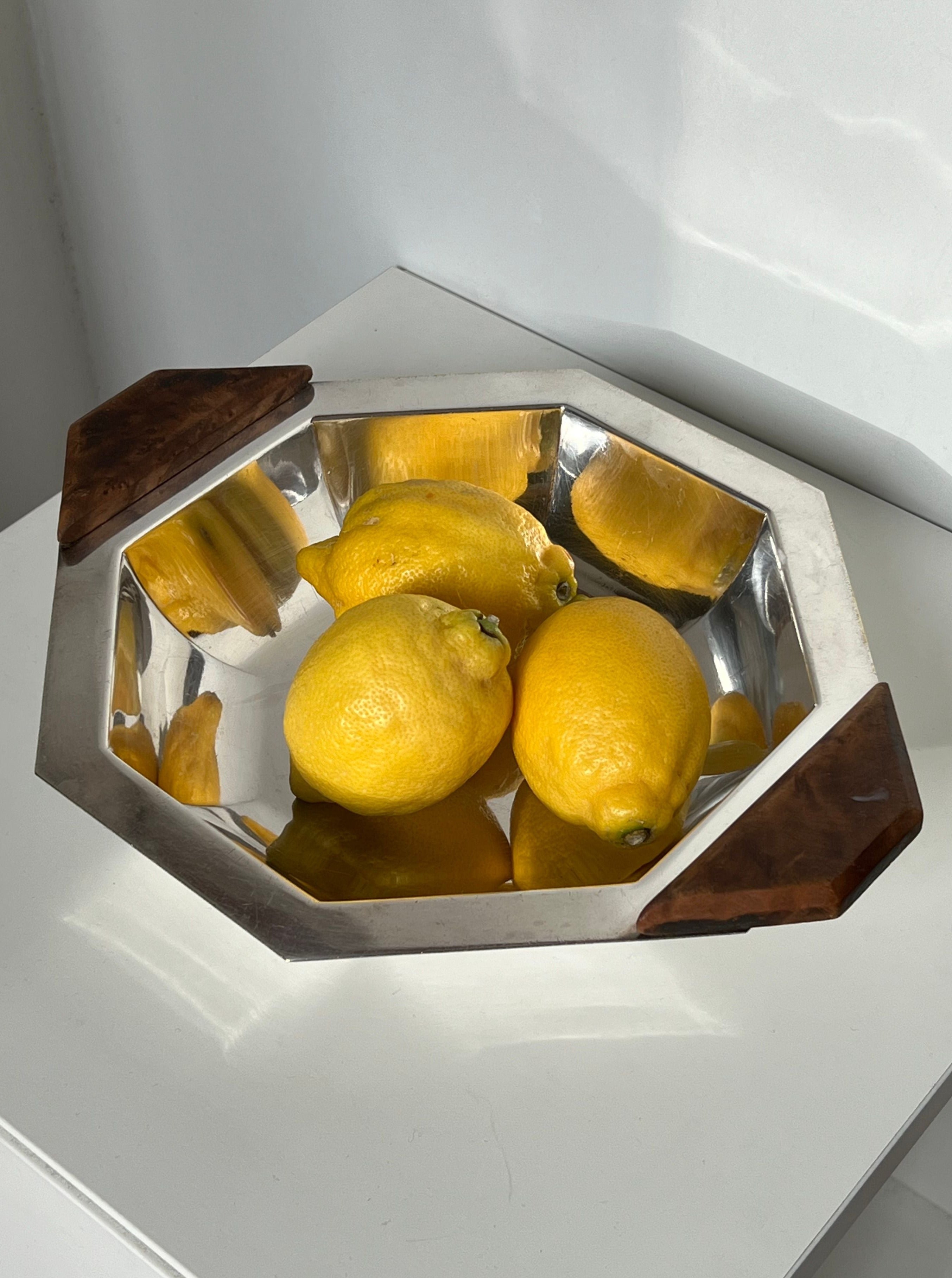 The Manushi Art Deco Decorative Dish, featuring a silver octagonal design and wooden handles, holds three yellow lemons on a white surface with light reflecting off both the dish and lemons.