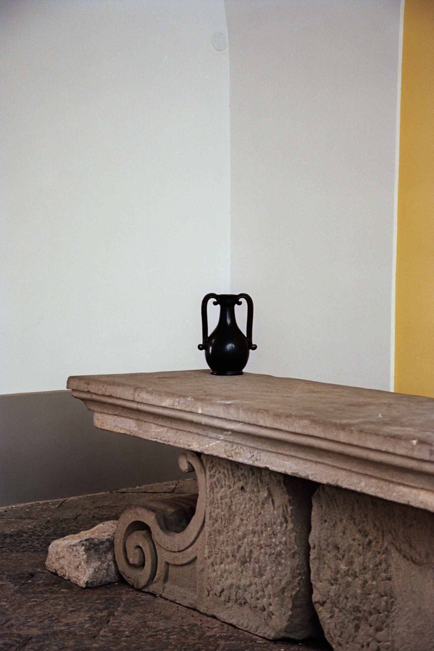 The Fiasca - Minimalist Vase in Ceramic by COSEINCORSO, a black terracotta piece with two handles, handmade in Italy, is displayed on an ornate stone table featuring carved scrolls, set against a plain white and yellow wall.