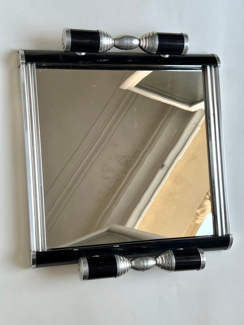 The Manushi Art Deco Mirrored Serving Tray features a rectangular design with a black and silver frame and cylindrical metallic accents on the top and bottom, reflecting elegant Art Deco style.