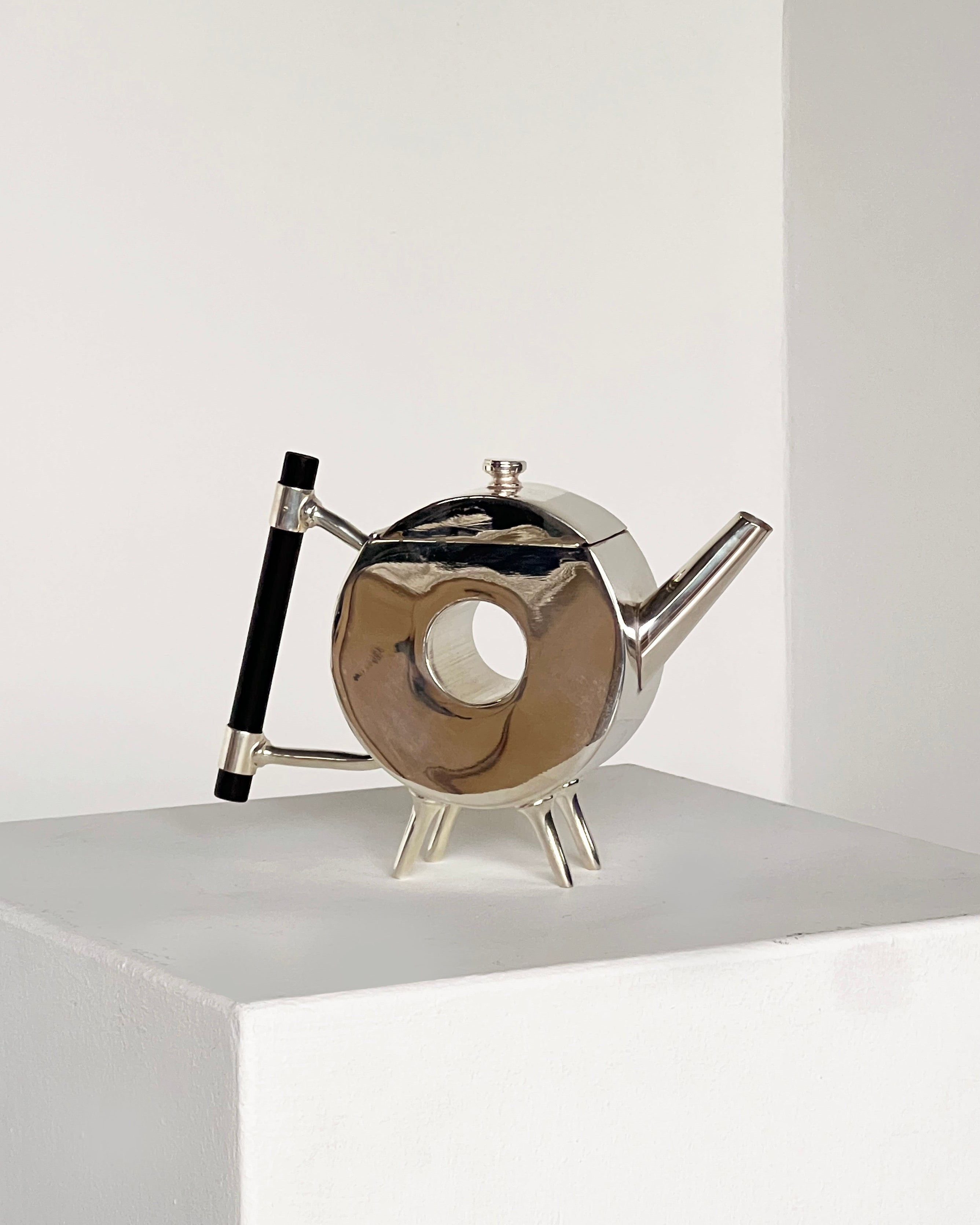 The Christopher Dresser Round Window Ornate Teapot by Porter Studio is a modern, silver-plated teapot with a circular body, black handle, angled spout, and three short legs displayed on a white pedestal against a plain background.