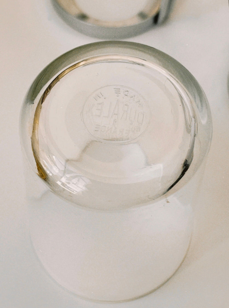 A close-up of an upside-down Manushi French Tea Glass With Stainless Steel Holder, featuring "DURALEX FRANCE" embossed on the bottom and reflected on a white surface—ideal for any refined tea glass set.