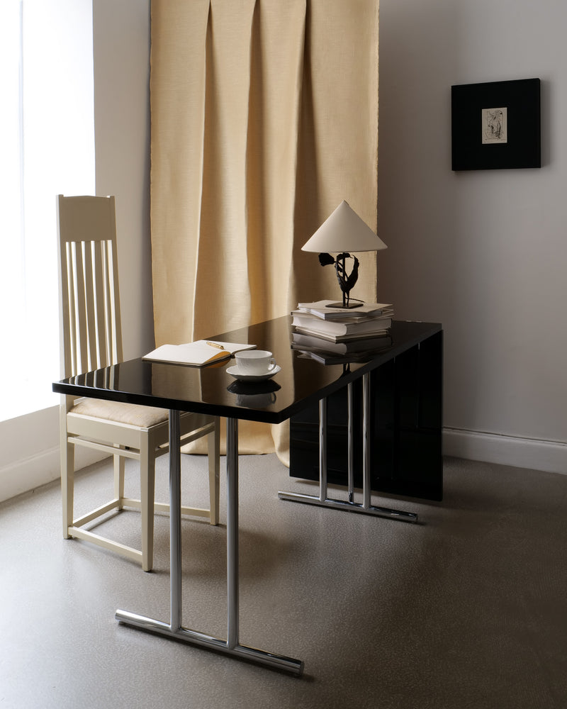 A minimalist workspace featuring the Lou Perou Extendable Table by Collection apart, paired with a cream chair, open notebook, cup and saucer, books, a lamp, beige curtain, and framed art highlights Eileen Gray's timeless elegance.