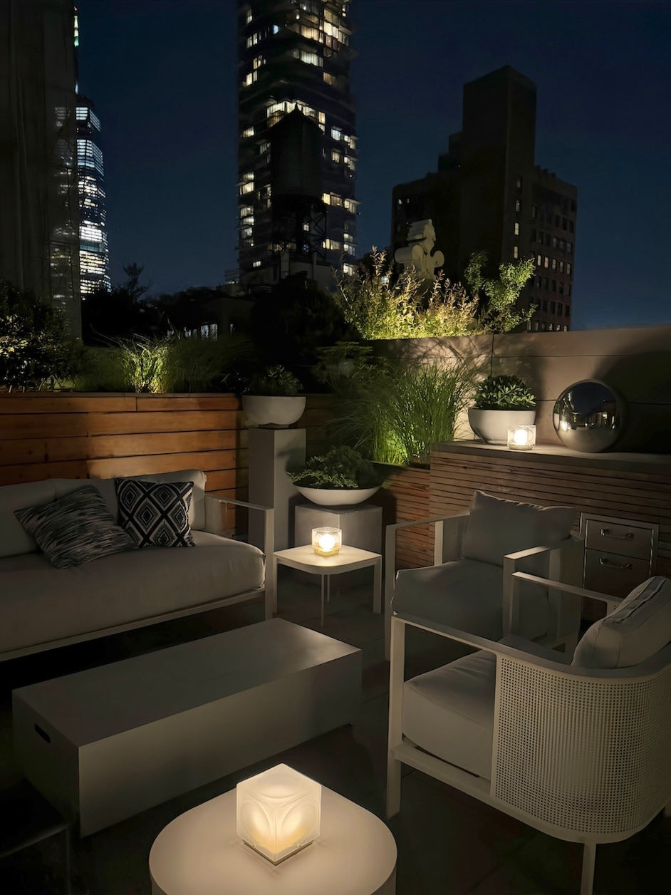 A modern rooftop patio at night features cozy seating, potted plants, and soft lighting from the Lee Broom Chant Portable Table Lamp, with tall city buildings illuminated in the background.