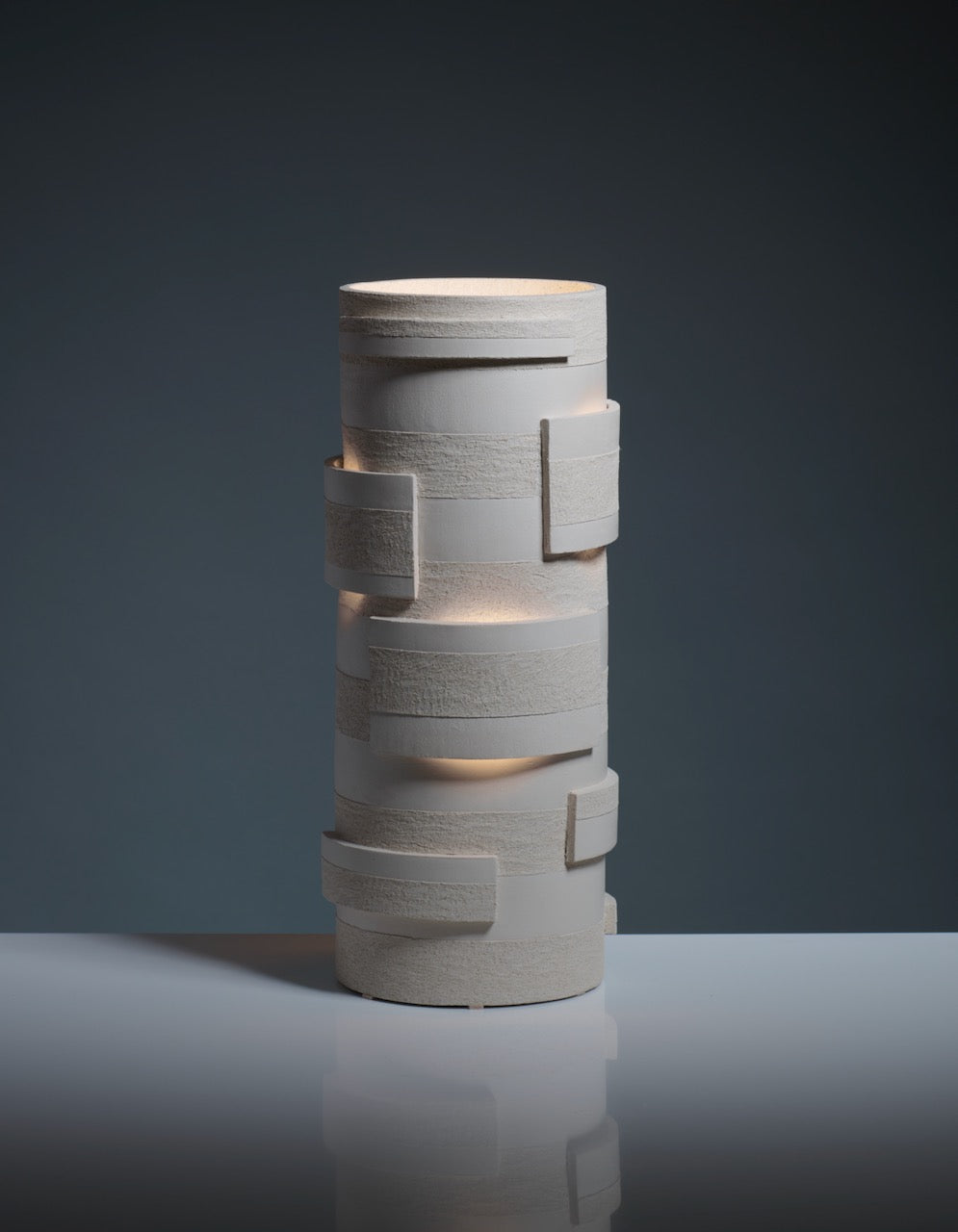 The "Bricks" Table Lamp by Katia Langeard features a modern cylindrical ceramic design with geometric layered rectangles, emitting soft light through gaps and shown on a reflective surface against a dark background.
