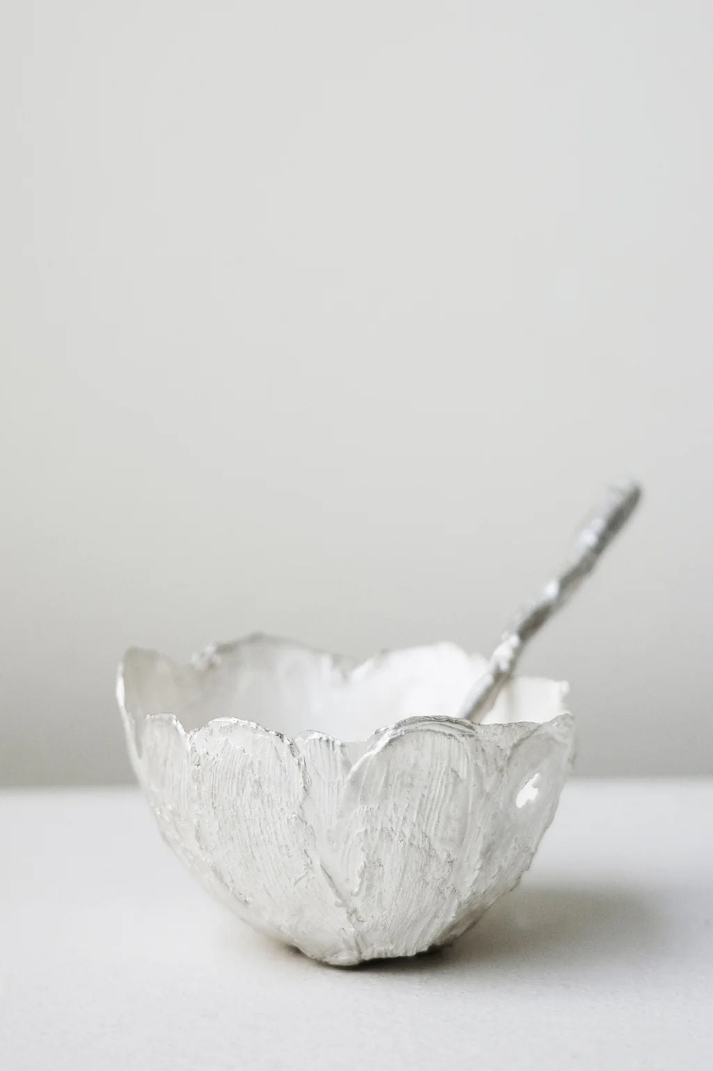 The L34 - Small Bowl in Silver by Completedworks, featuring a scalloped edge and textured finish, rests with its matching spoon on a flat surface against a light background.
