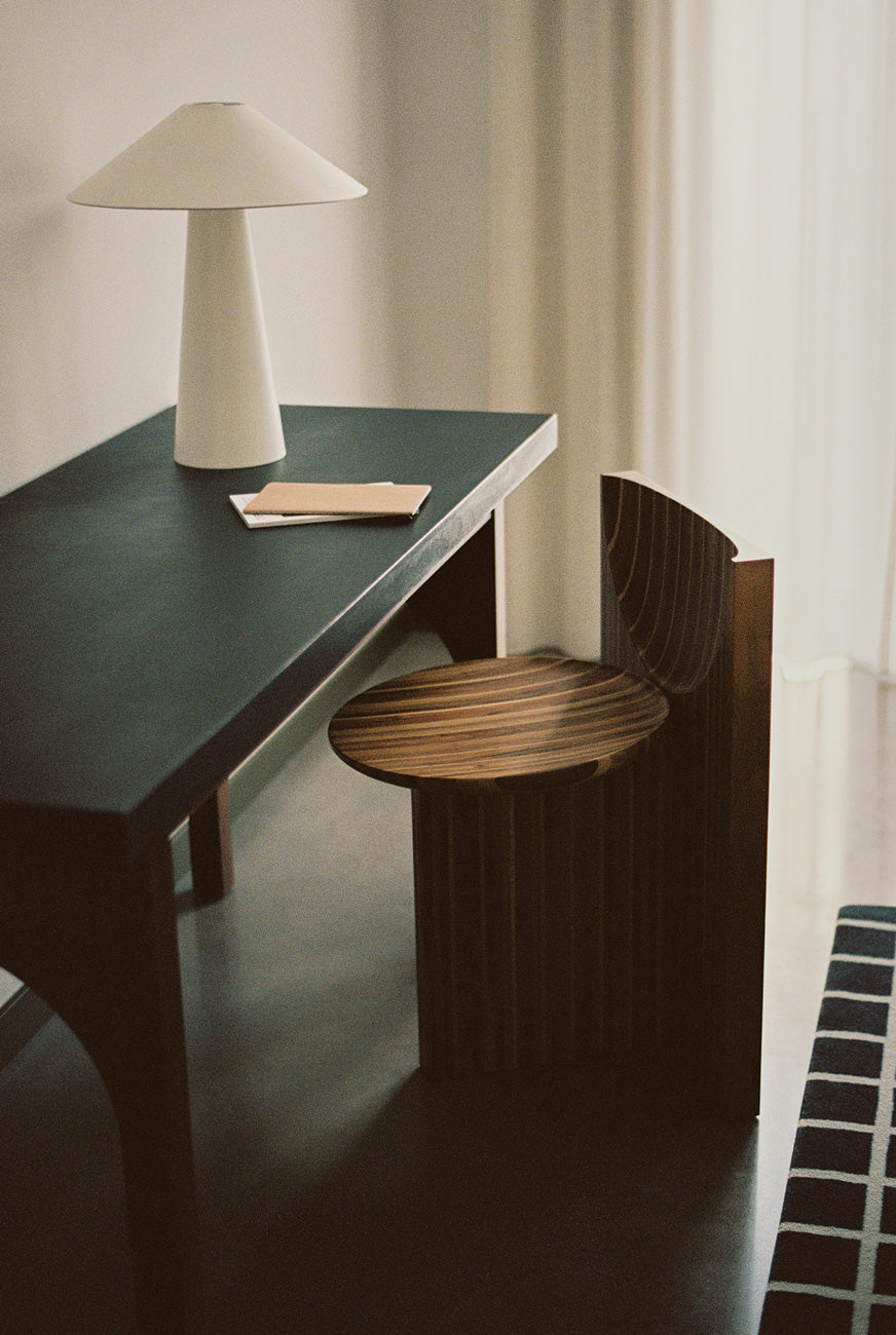 A minimalist interior features a sleek black desk complemented by a modern white lamp. The highlight of the room is the Vaga Chair from Rosana Sousa, crafted from solid walnut and oak wood, showcasing exquisite Portuguese woodworking. A folded paper sits nearby as light filters through the curtain, creating a serene atmosphere.