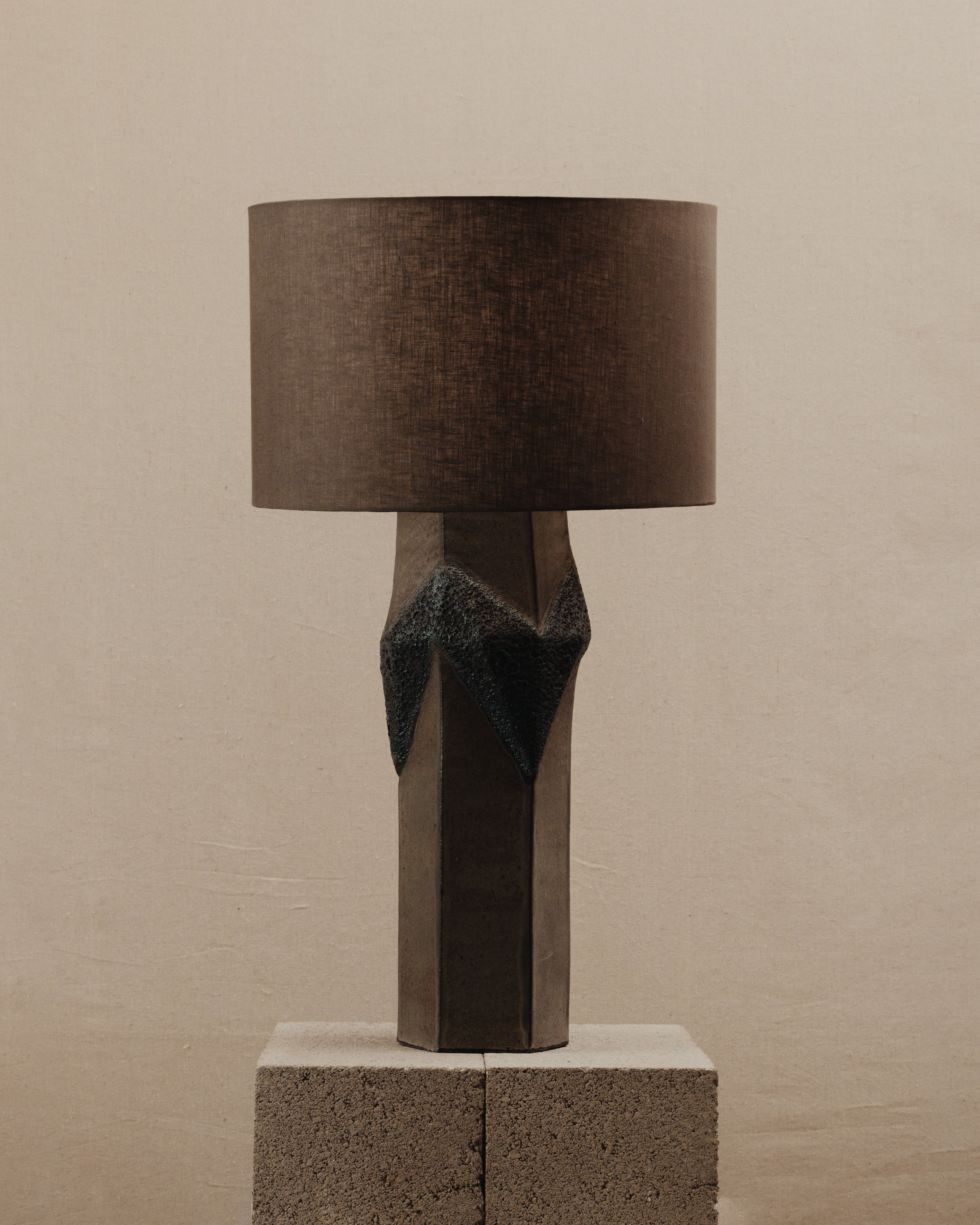 The Warp Table Lamp by JS/WRKSHP features a geometric ceramic base with a dark textured accent and a brown cylindrical shade, shown atop a rough concrete block against a plain beige backdrop.